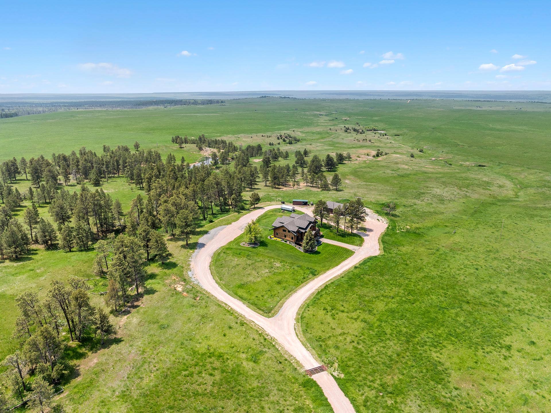 Aerial view of a large house at the end of a curved gravel driveway, surrounded by green fields and scattered trees.