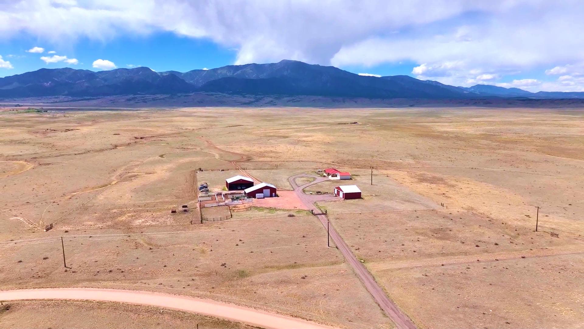 Remote farmstead in a dry plain with red-roofed buildings and a dirt road, mountains in the distance.