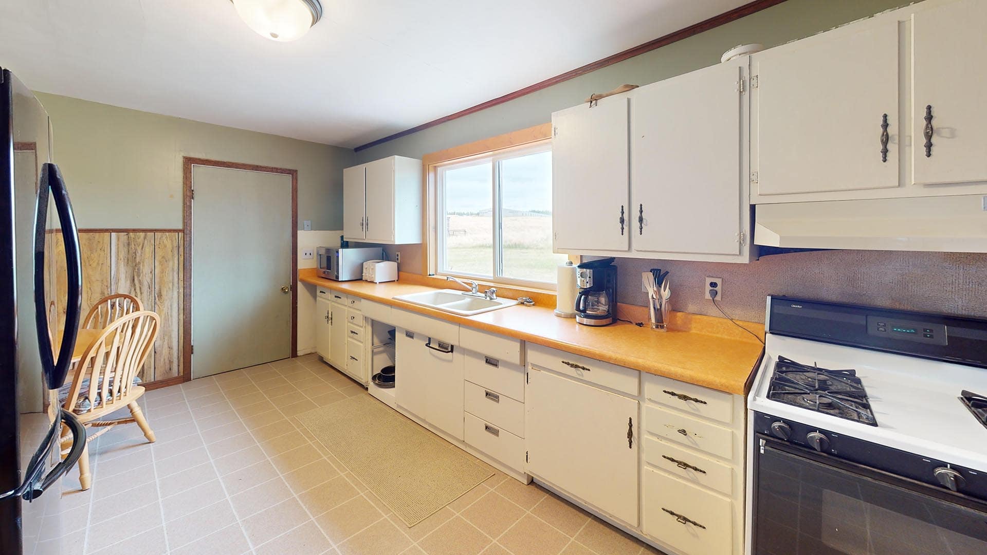 Bright kitchen with white cabinets, beige countertops, and a large window over the sink receding into a rural view.