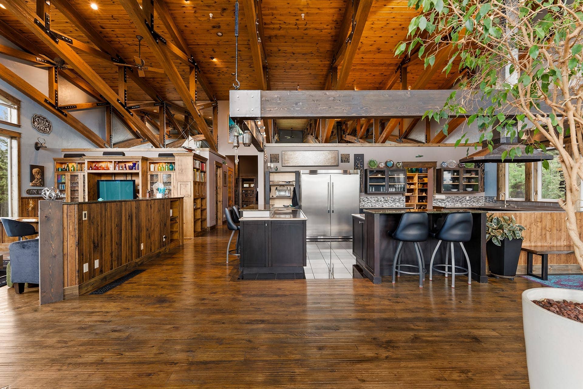 Open-concept kitchen with dark wood island, stainless fridge, and wooden beams, large windows, and indoor tree fills the space.