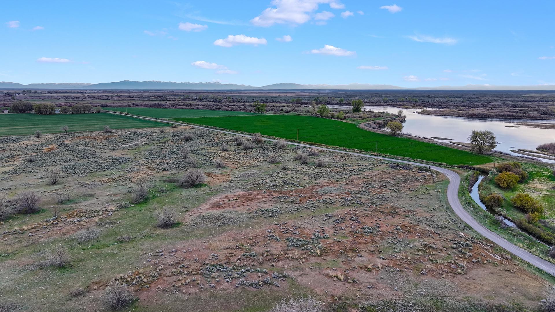 Aerial view of green farmland beside a winding river, with a dirt road and distant mountains under a blue sky.