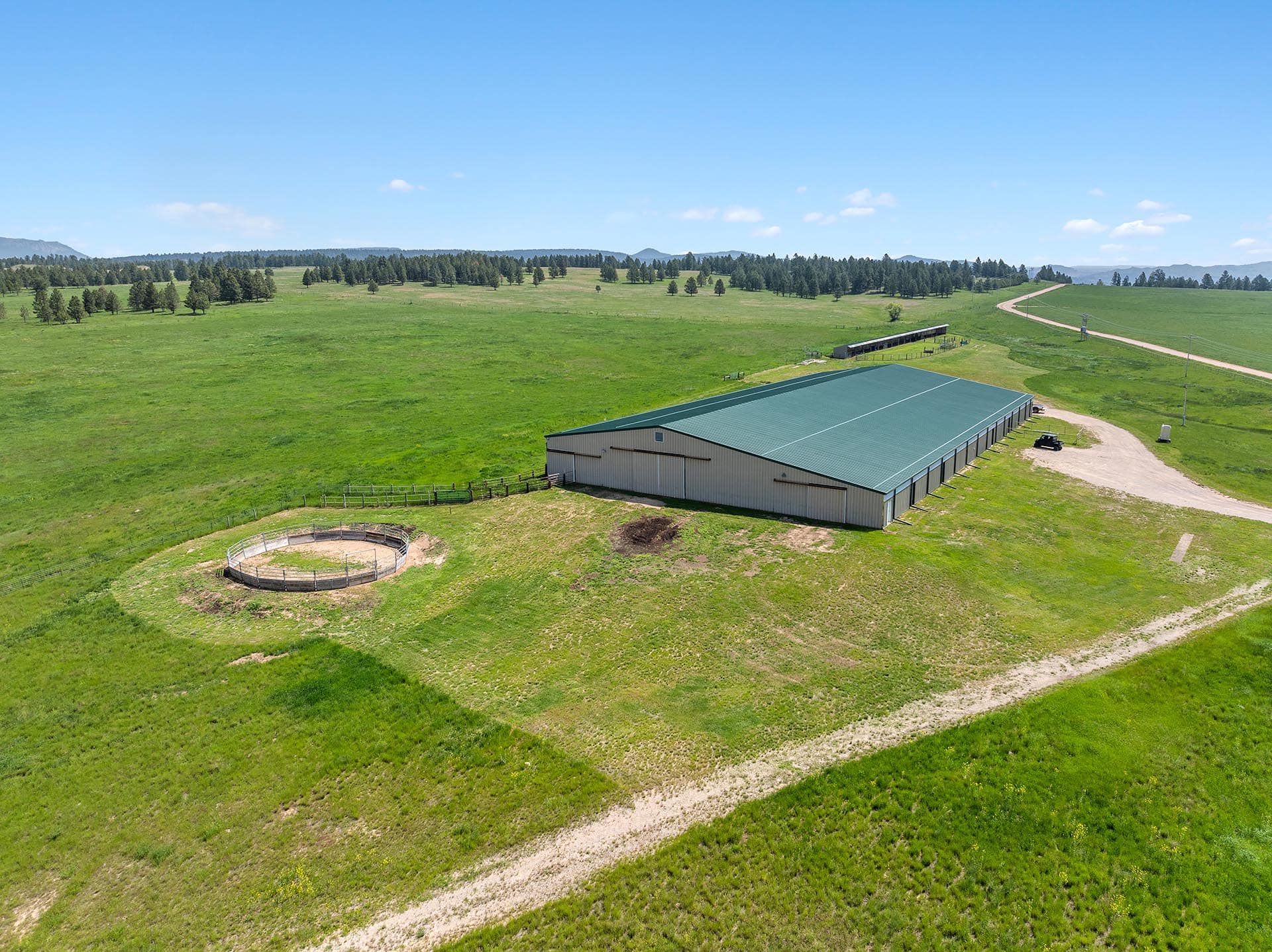 A large metal-roofed barn with a circular outdoor pen nearby on a green field, dirt road in foreground.