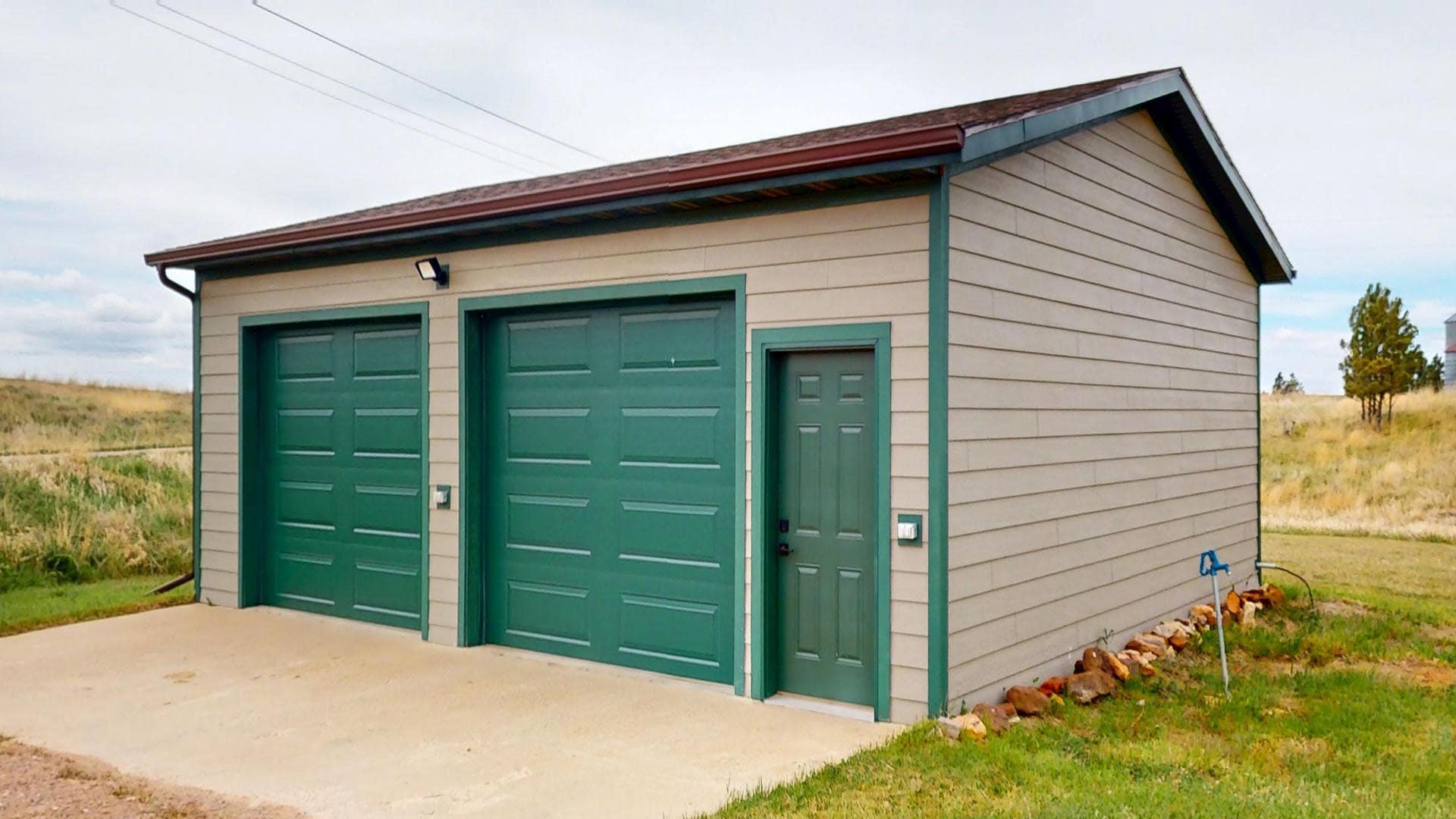 Small beige garage with two green doors and a side entrance in a rural yard, beige siding and green trim near an open field