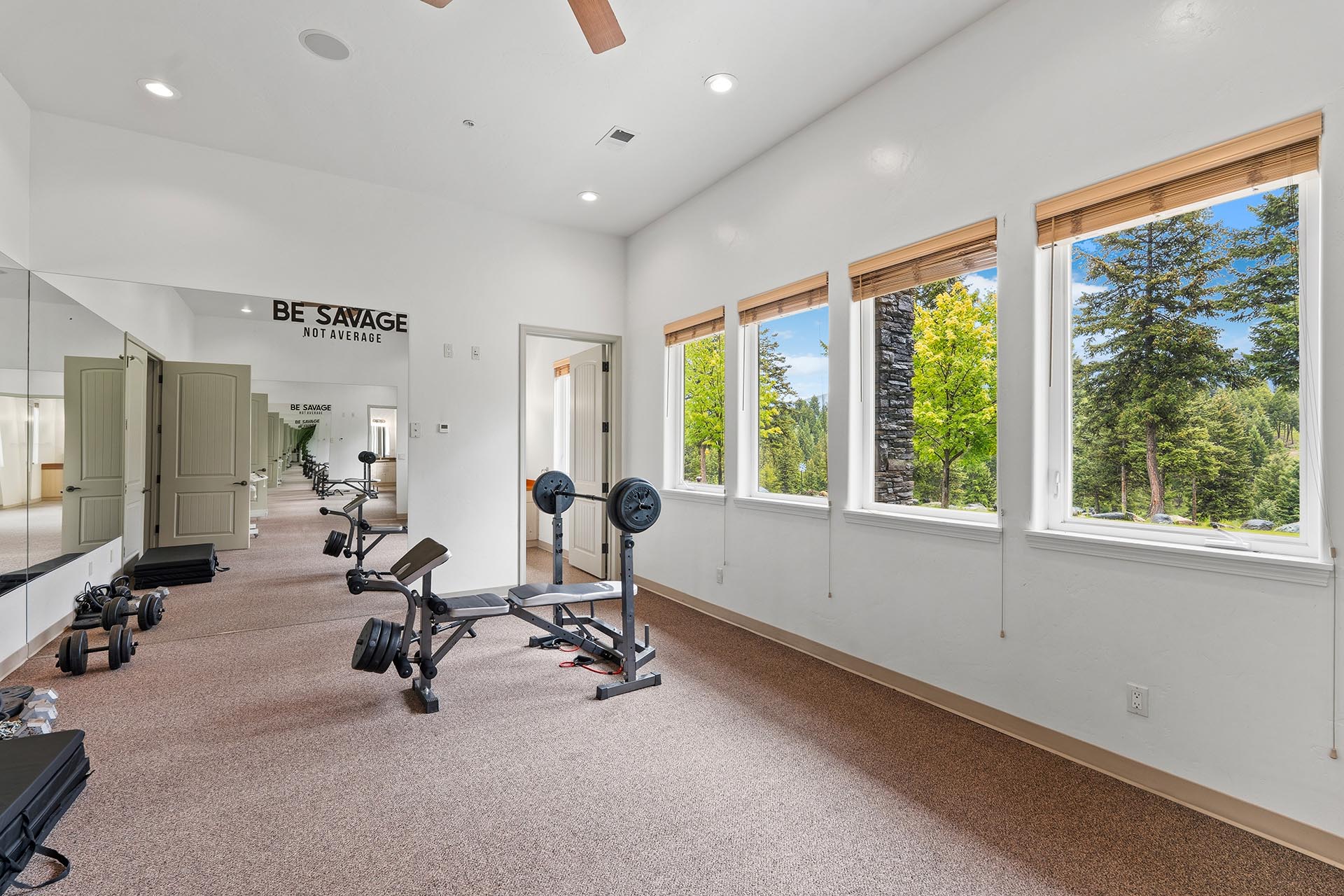 Home gym with a weight bench, dumbbells, and mirrors, large windows showing trees outside.
