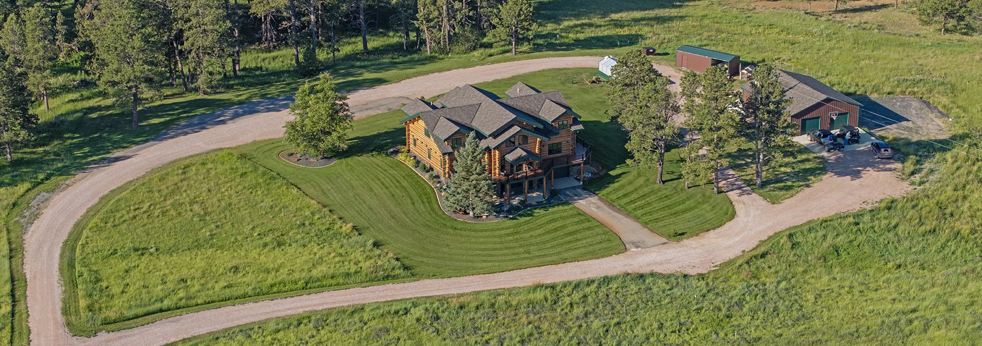 Aerial view of a log home with a curved gravel driveway and a neatly mowed lawn surrounded by fields and trees, in a rural setting.
