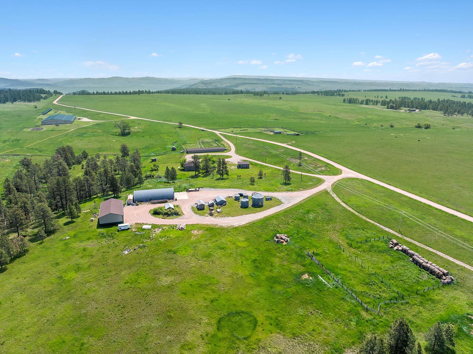 Aerial view of a rural farmstead with several buildings, a curved dirt road, and expansive green pastures under a blue sky.