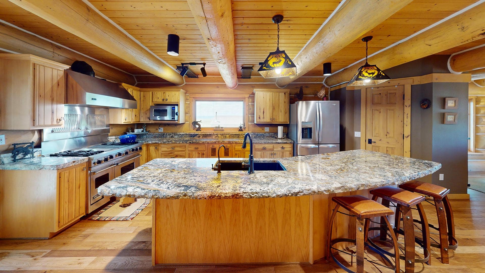 Rustic wooden kitchen with a large granite island, stainless steel appliances, and exposed ceiling beams in a cabin-style home.