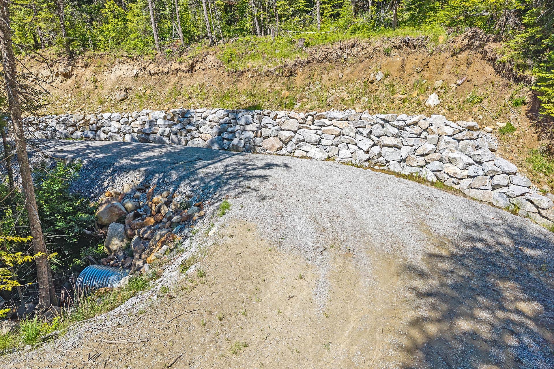 Stone retaining wall along a gravel road, supporting a dirt hillside with trees behind it. A blue corrugated drainage pipe is visible at the bottom left.