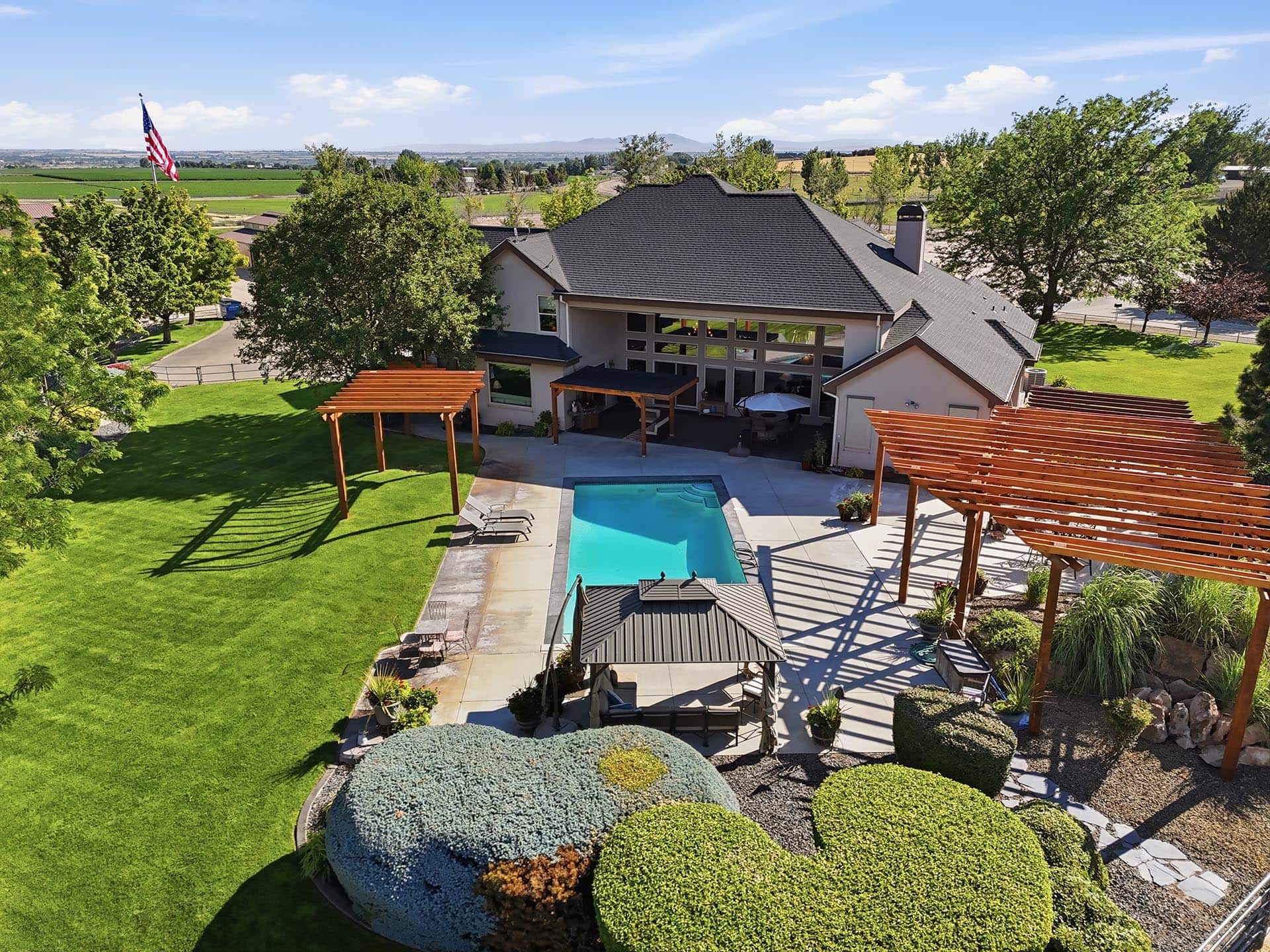Aerial view of a modern house with a rectangular pool, wooden pergolas, green lawn, and manicured shrubs in a spacious backyard.