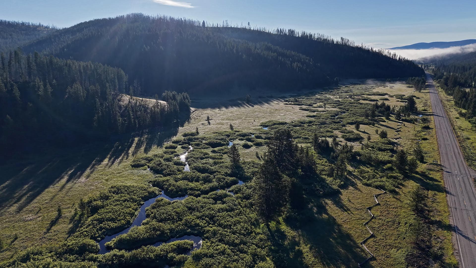 Aerial view of a grassy valley with a winding stream, forested hills on the left, and a long road along the right edge.