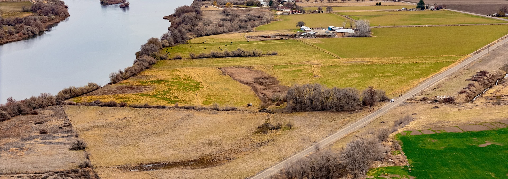 Aerial view of rural farmland along a river: patchwork fields, a road cutting diagonally, and farm buildings in the distance, with trees lining the riverbank.
