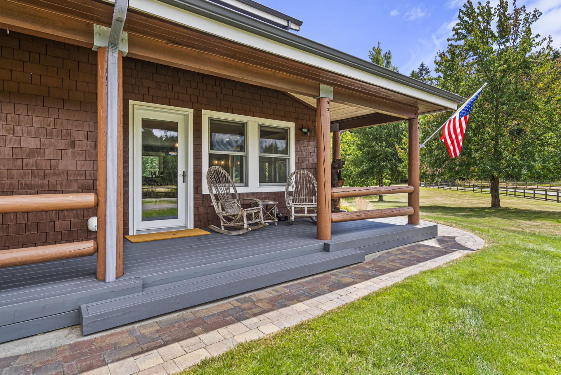 Front porch of a brown wooden house with two wicker rocking chairs, a white door, and an American flag waving beside trees in a sunny yard.