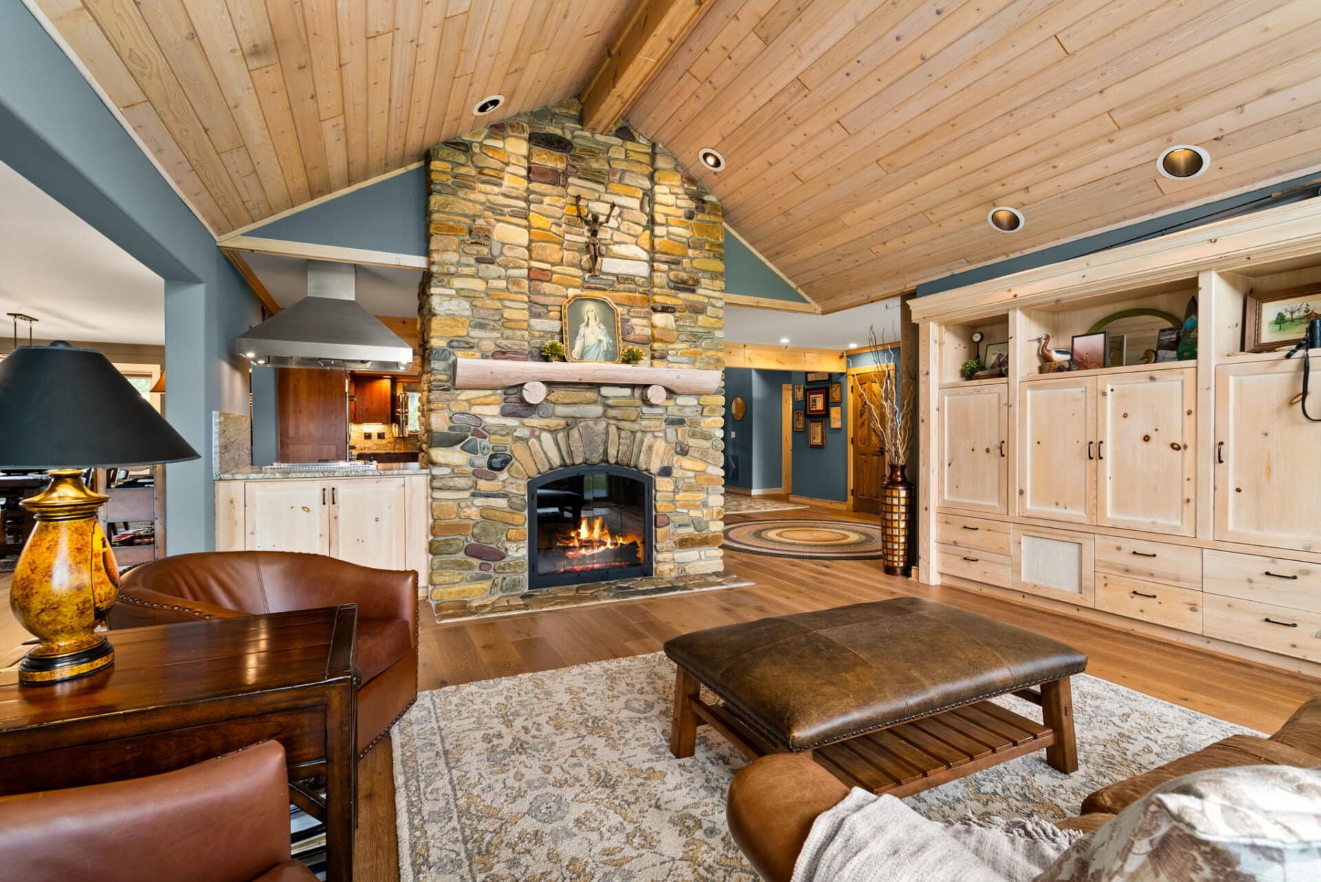 Cozy living room with a tall stone fireplace, vaulted wooden ceiling, and built-in light wood cabinets along the wall.