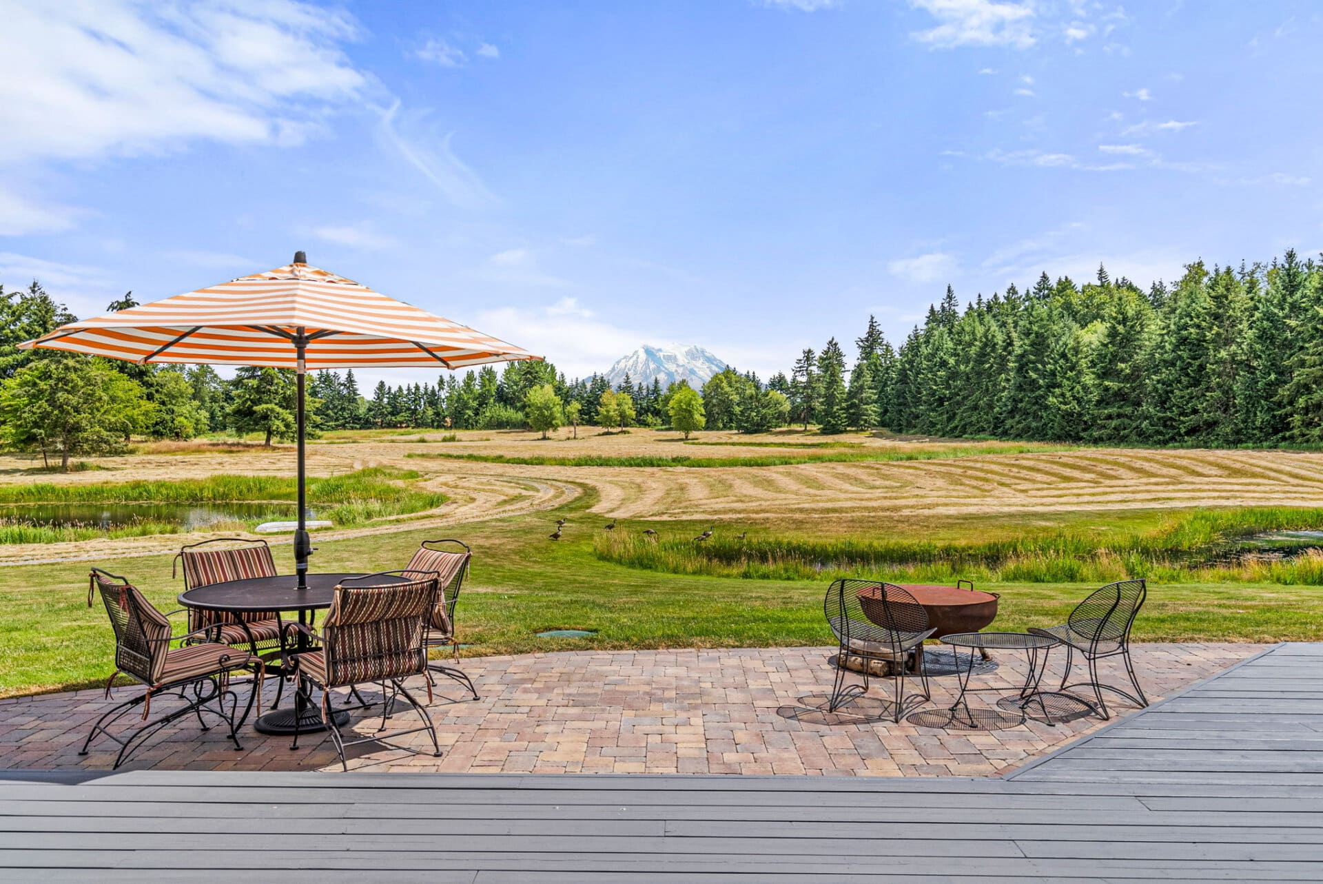 Outdoor patio with a striped orange-and-white umbrella over a round table and wicker chairs, overlooking a grassy field and distant mountains.
