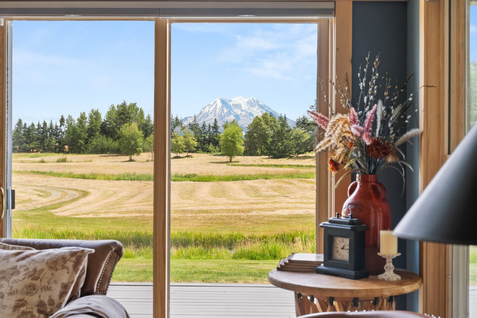 Living room with a large window showing a field, trees, and distant snow-capped mountains; a red vase with dried flowers sits on a round table nearby.