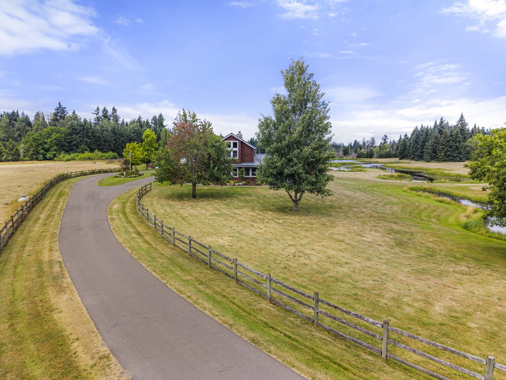 Red brick house with a pointed roof framed by trees, a curved driveway, and a wooden fence on a grassy rural lawn under a blue sky.