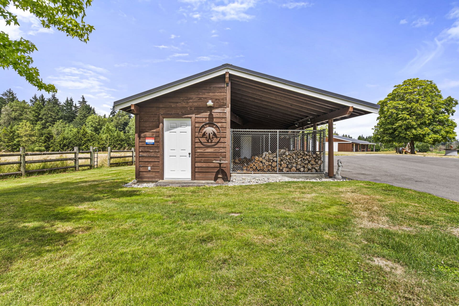 Brown wooden shed with white door, open-sided log storage under a pitched roof, and a chain-link enclosure of stacked firewood beside it on a sunny day