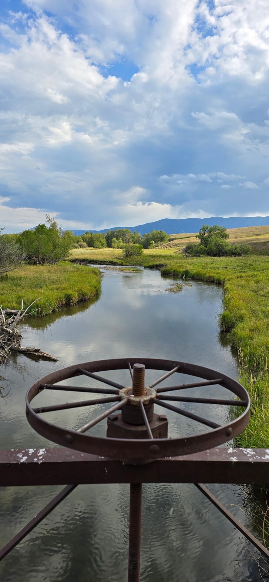 Rusty metal water wheel mounted on a railing over a calm river winding through grassy plains with trees and distant hills under a cloudy sky.