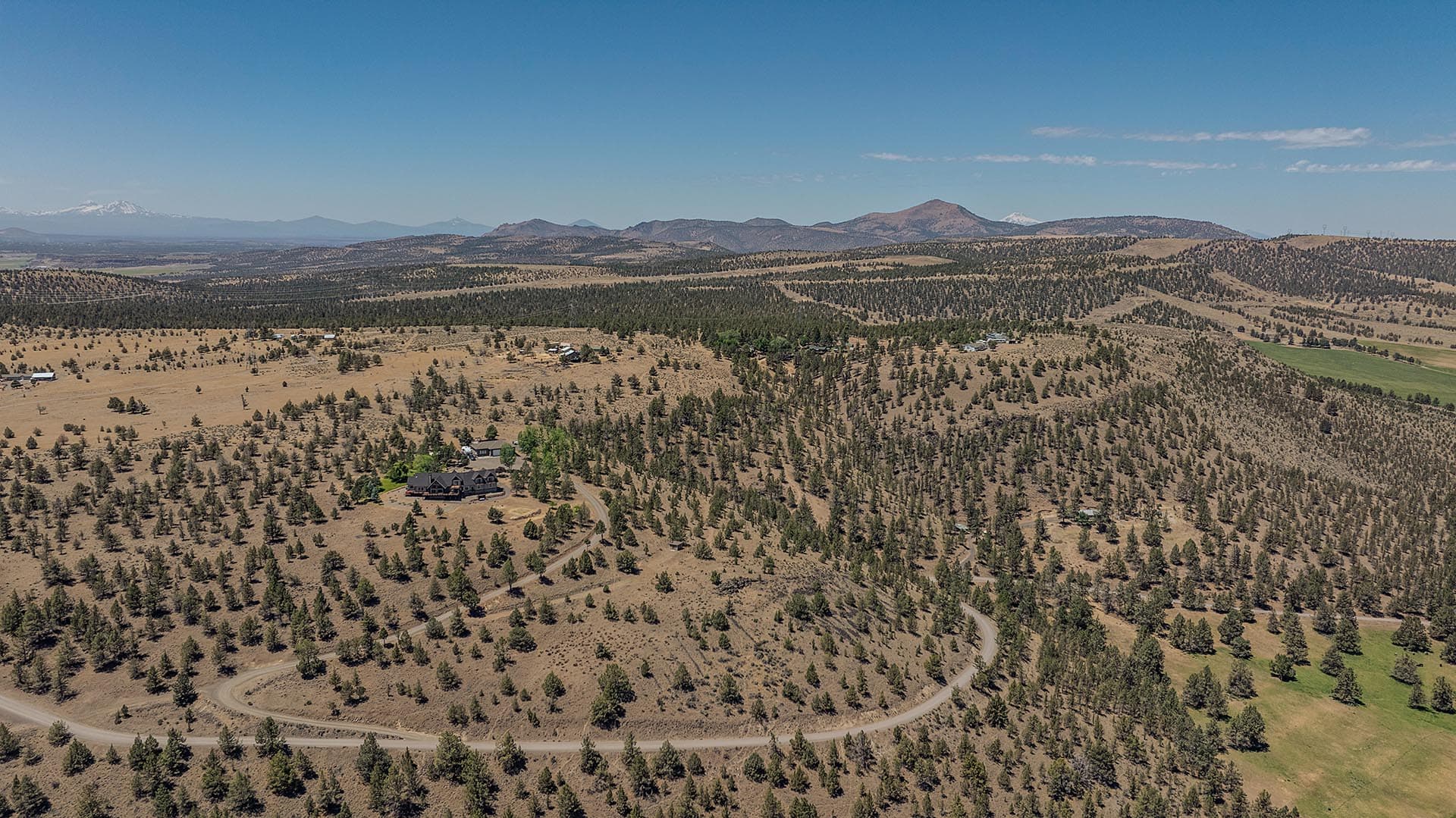 Aerial view of a dry, pine-dotted hillside with a winding road leading to a large house and distant mountains under a clear blue sky,