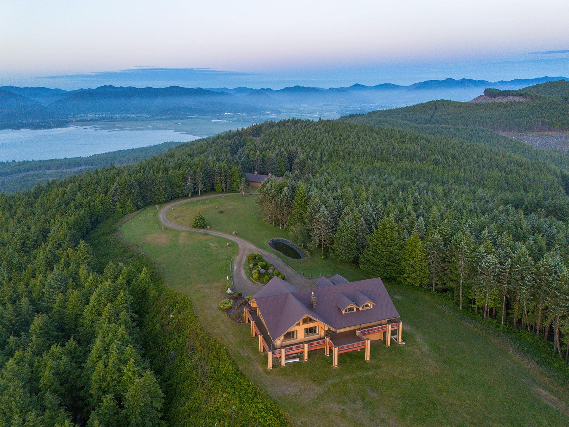 Aerial view of a large wooden lodge surrounded by dense forest, with a lake on the left and distant mountains on the horizon.