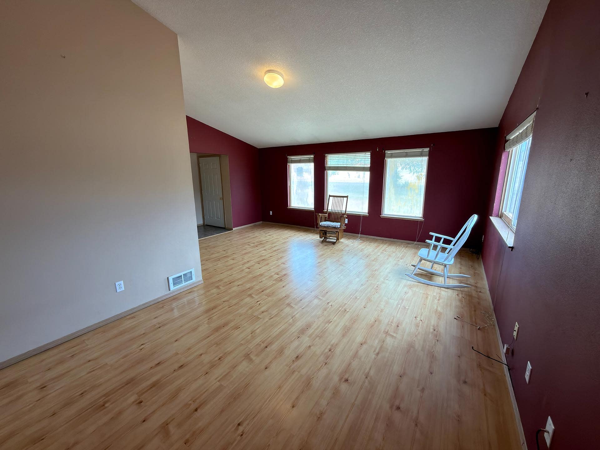 Empty living room with red accent walls, light wood floor, and three windows letting in daylight; a couple of chairs near the windows.