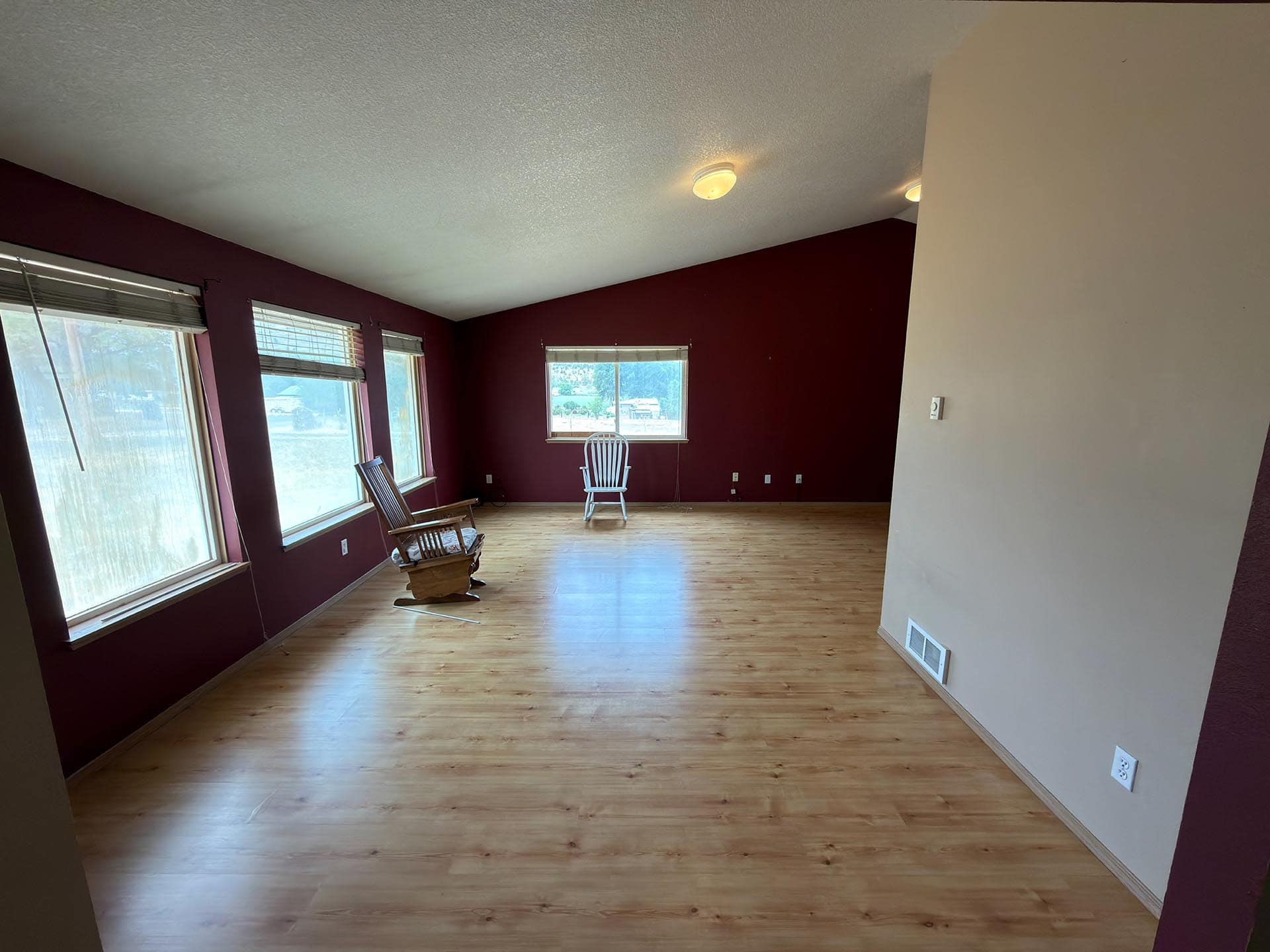 Living room with burgundy walls and light wood floors, three large windows along one wall, and a few wooden chairs scattered around.