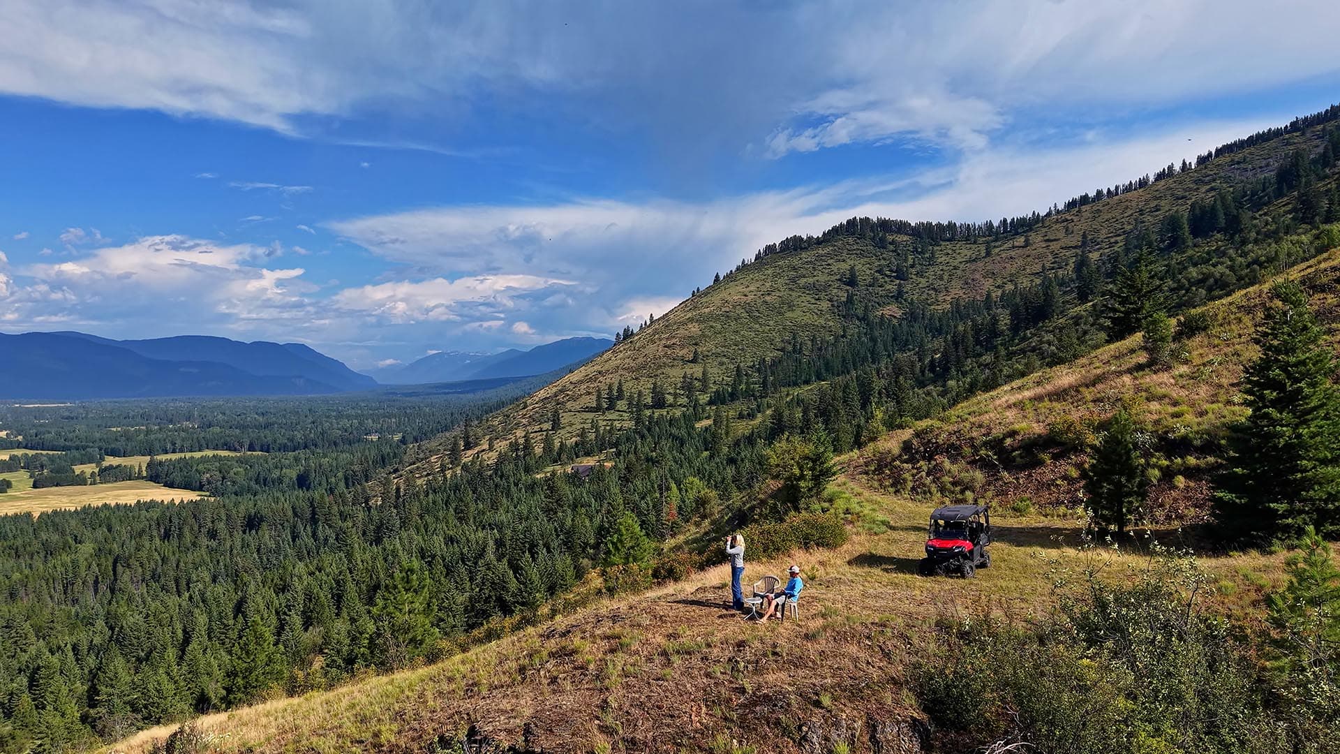 Scenic hillside overlooking a valley with forests and distant mountains; two people sit and a person stands nearby while a red ATV rests on the slope.