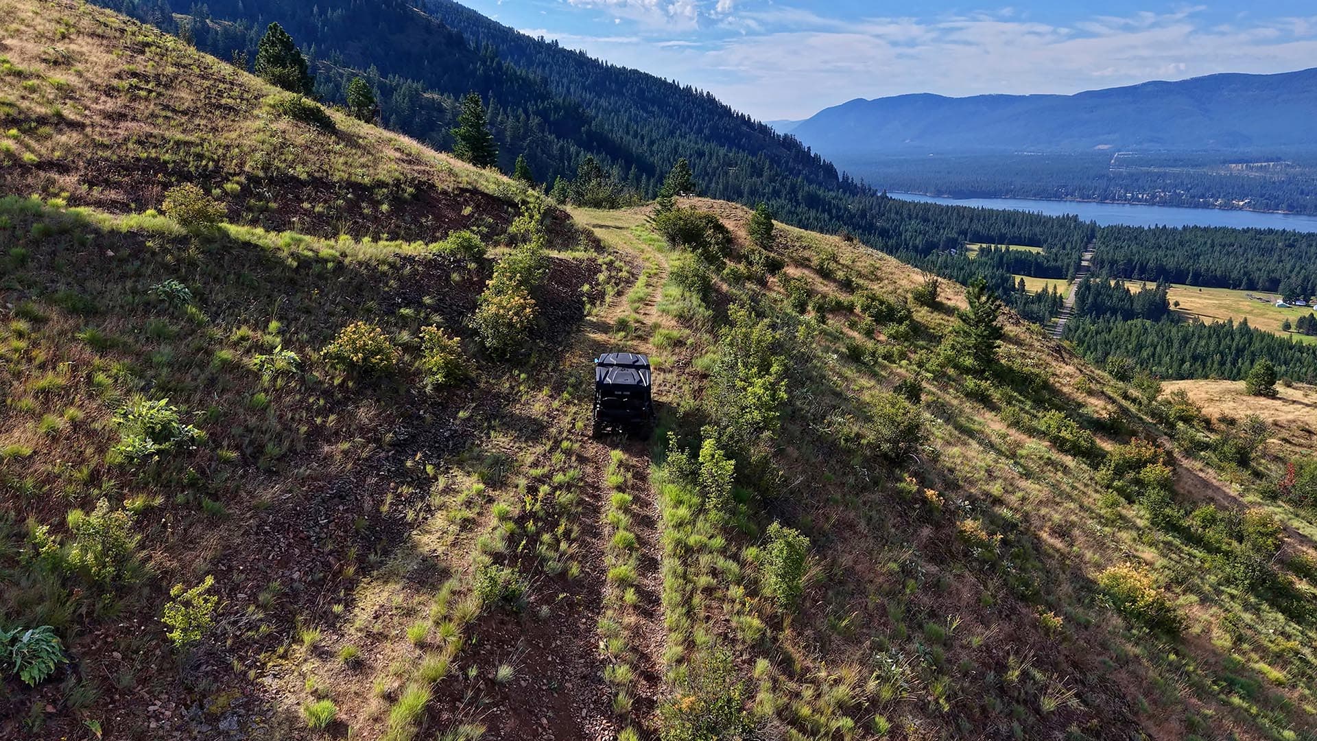 Off-road vehicle on a rocky hillside trail with pine trees and distant lake and mountains in the background.
