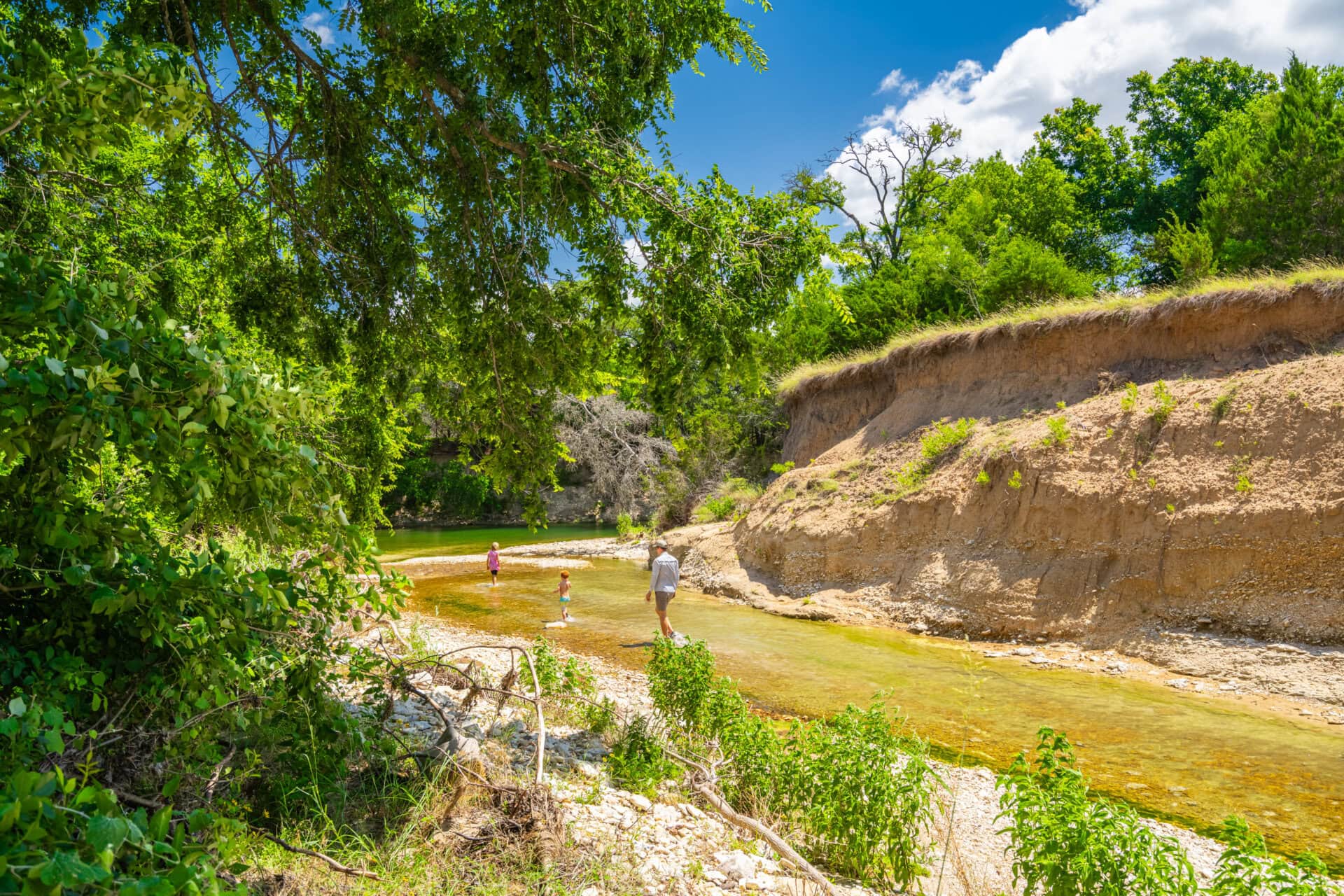 Children wading in a shallow river along a sandy bank, framed by overhanging green trees and a steep earthen cliff under a bright blue sky.