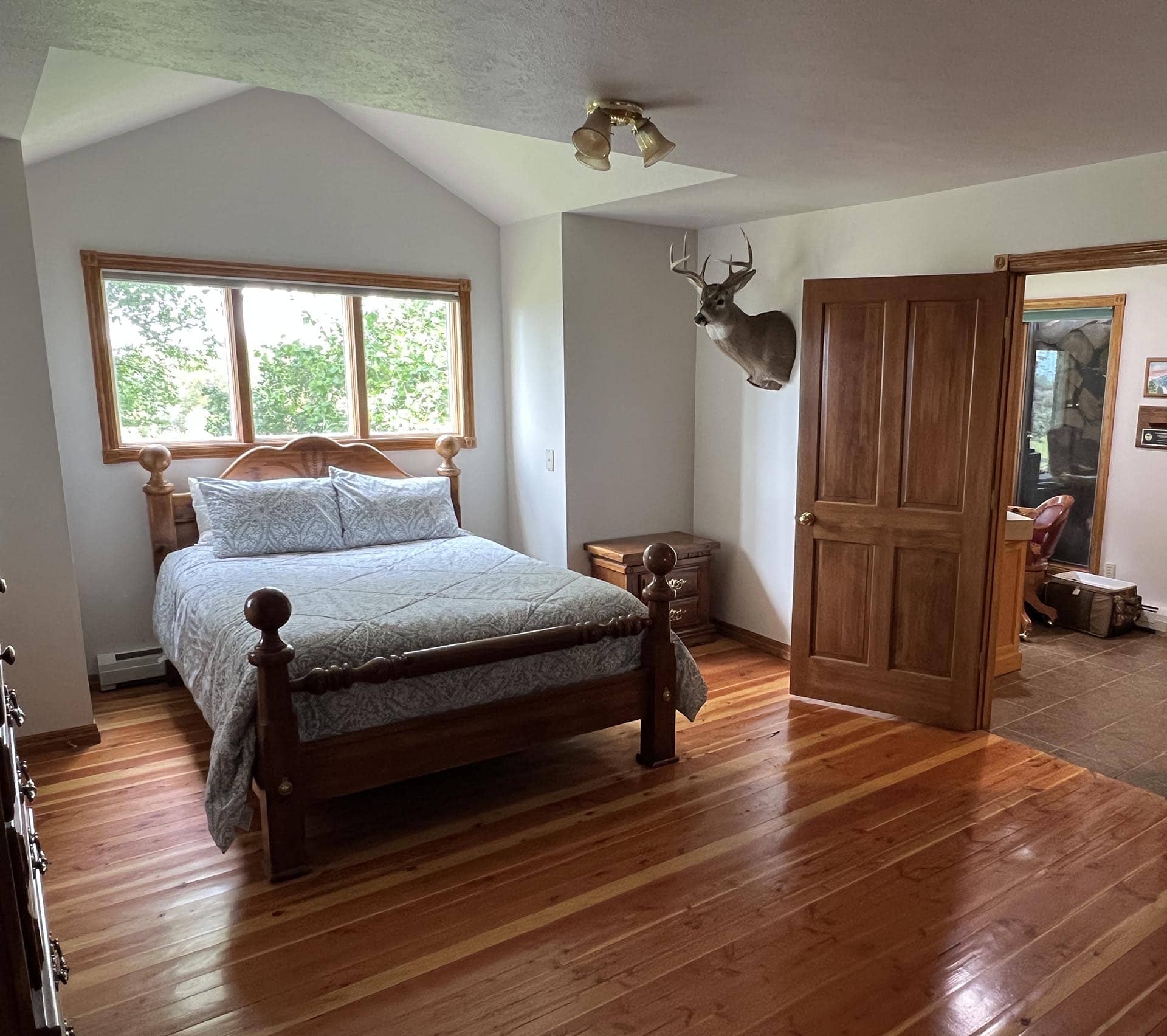Bedroom with a wooden four‑poster bed, blue bedding, and a mounted deer head on the wall by a large window. Sunlight through the trees outside.