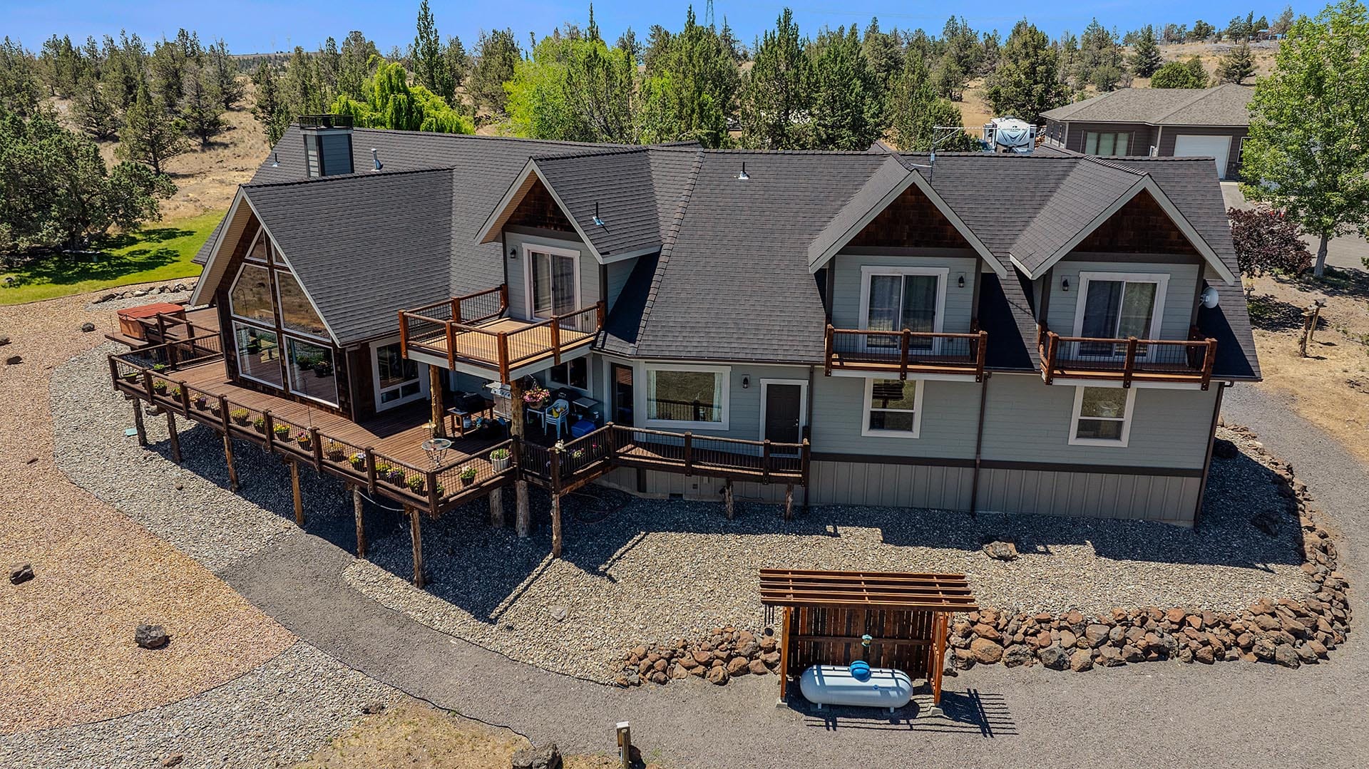 Aerial view of a two-story lakeside home with multiple wooden decks, pebble beach, and surrounding trees.