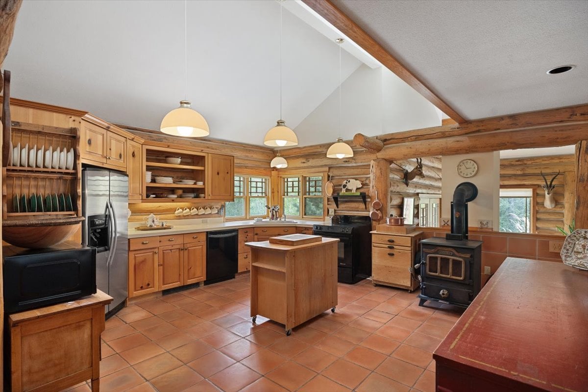 Rustic log-cabin kitchen with wooden cabinets, a central movable island, pendant lights, and a wood-burning stove along a tiled floor.