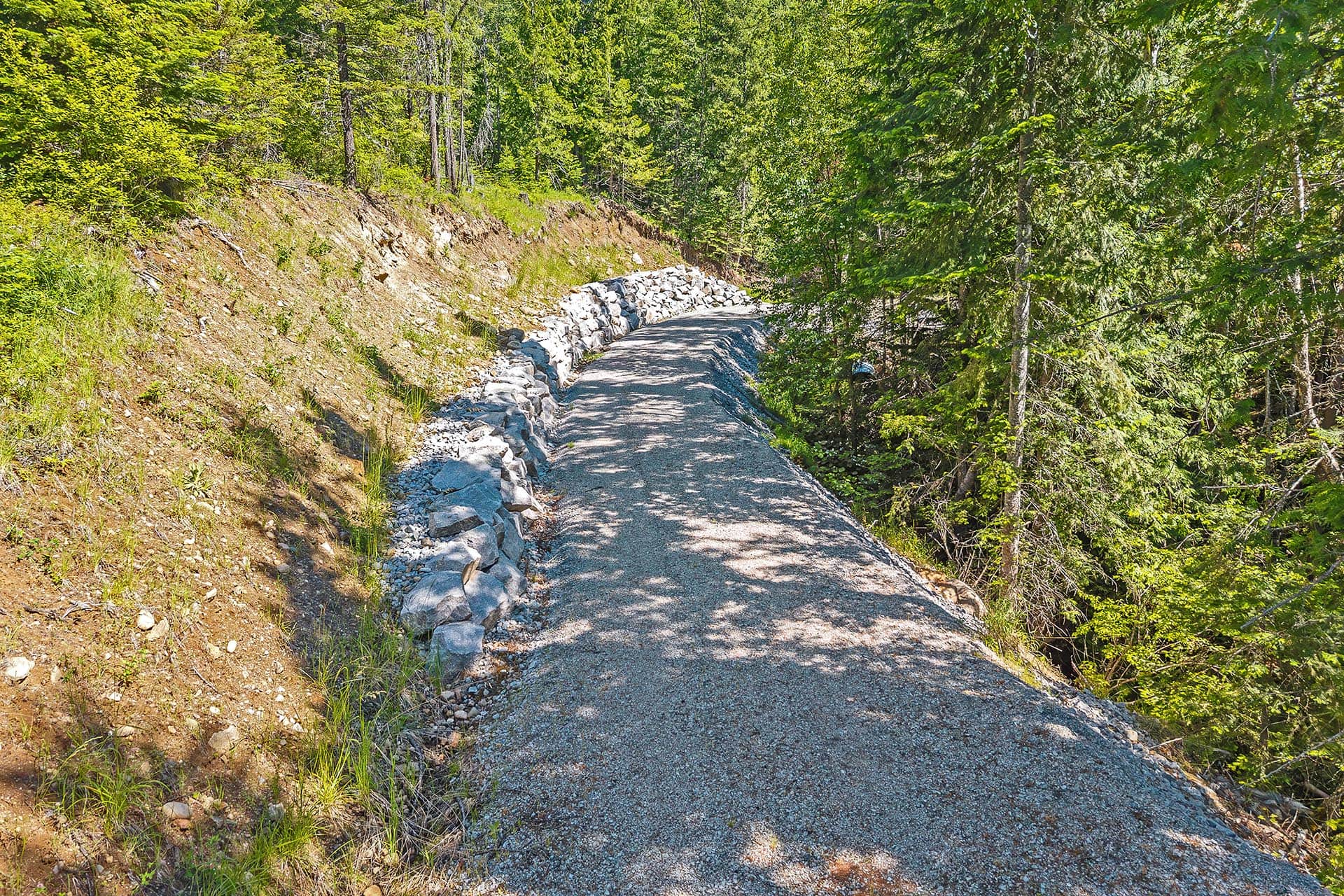 Gravel trail winding through a wooded hillside with a stone retaining wall on the left.