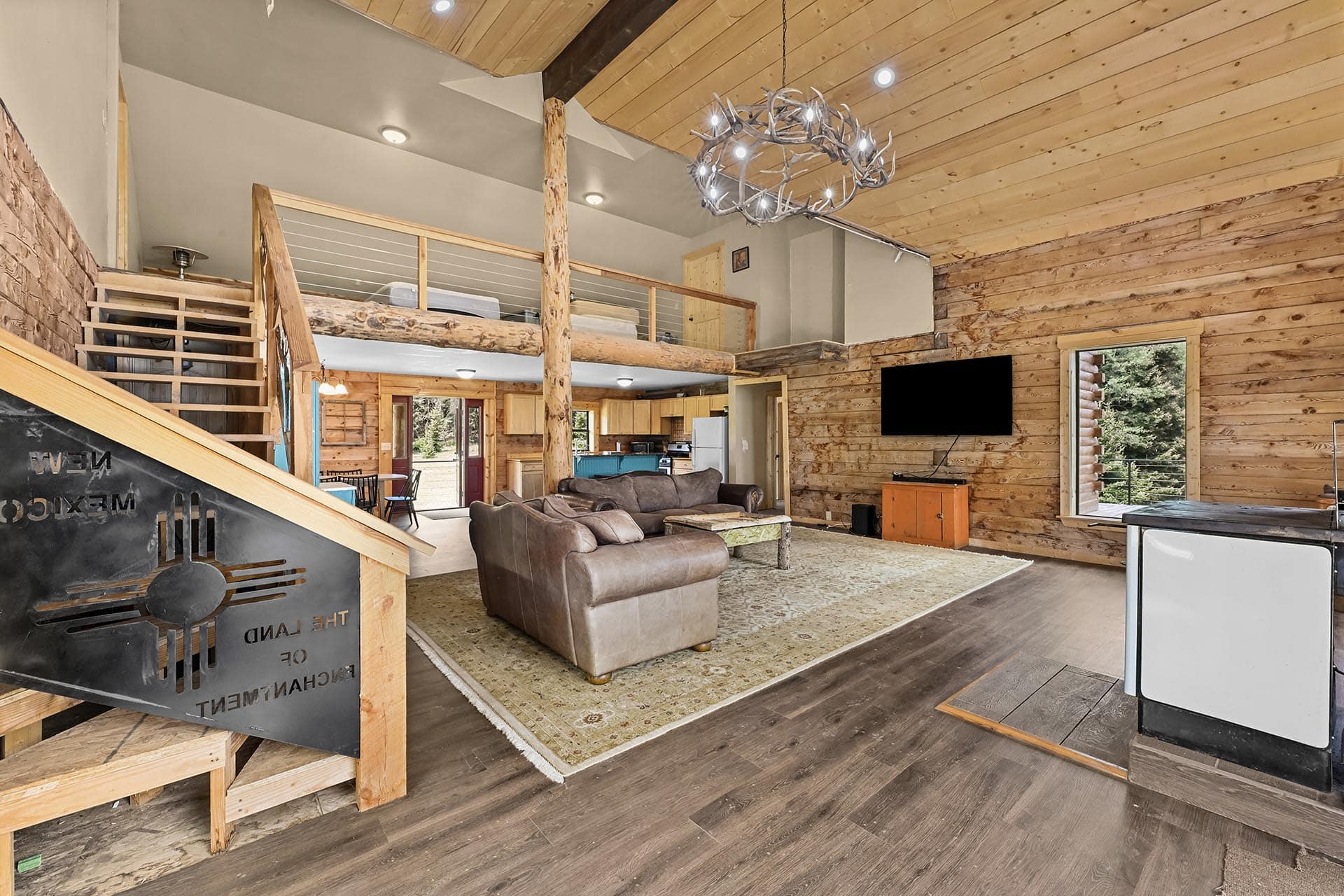 Open-concept log cabin living area with exposed wood walls, a mezzanine balcony, antler chandelier, and a leather sectional facing a wall-mounted TV.