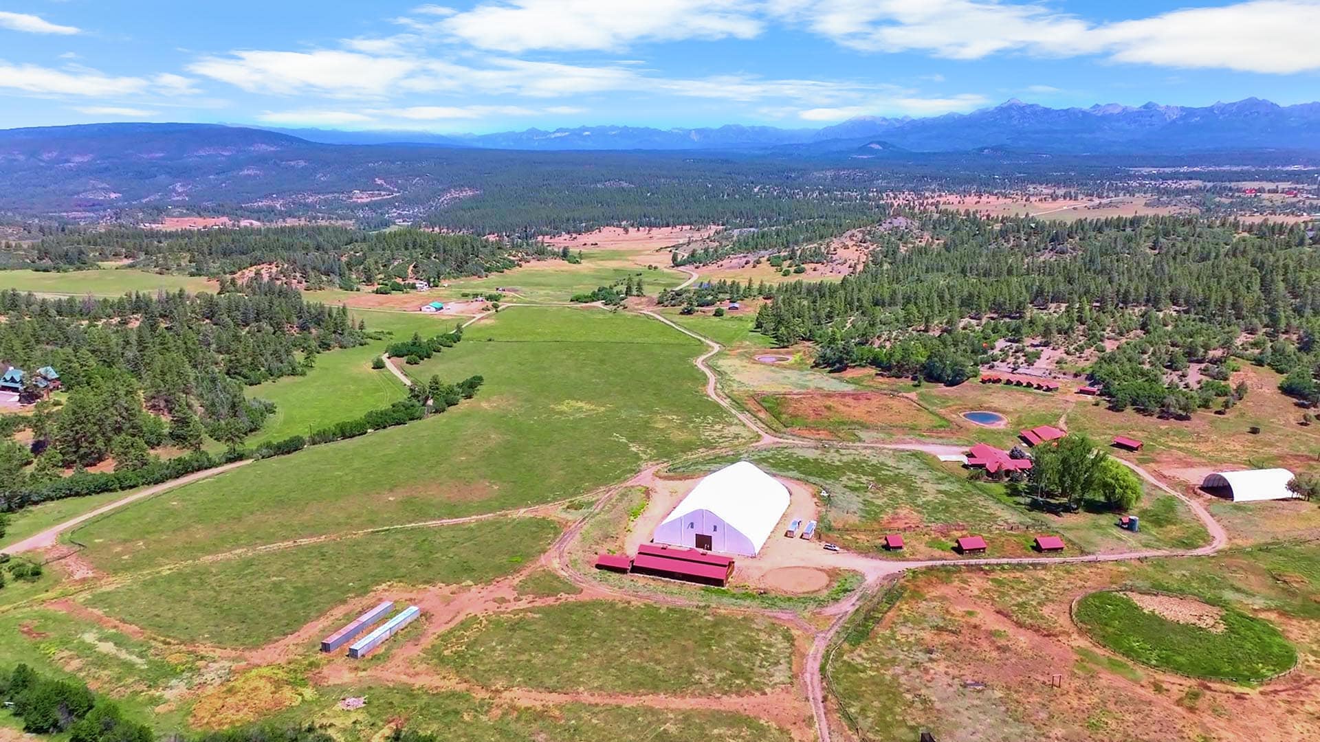 Aerial view of a rural farm with red barns, green fields, and distant mountains under a blue sky.