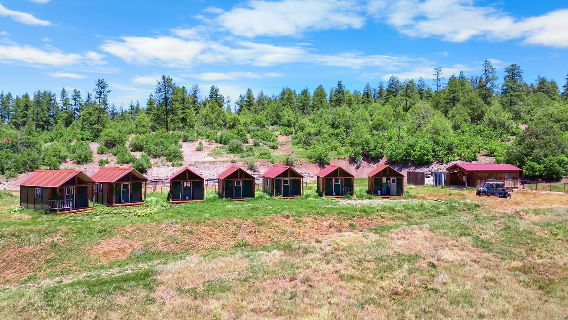 Row of red-roofed cabins at a campsite, green grass in front and a forested hill behind under a blue sky.