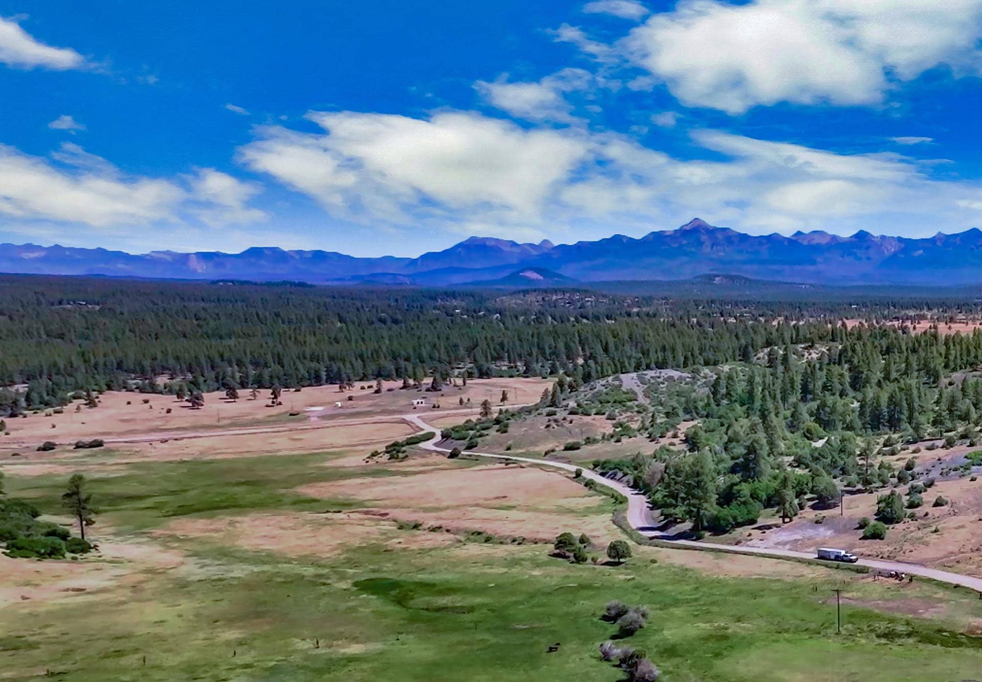 Wide landscape view of rolling fields and a forest with distant mountains under a blue sky with scattered clouds, a winding road and a white vehicle bottom-right.