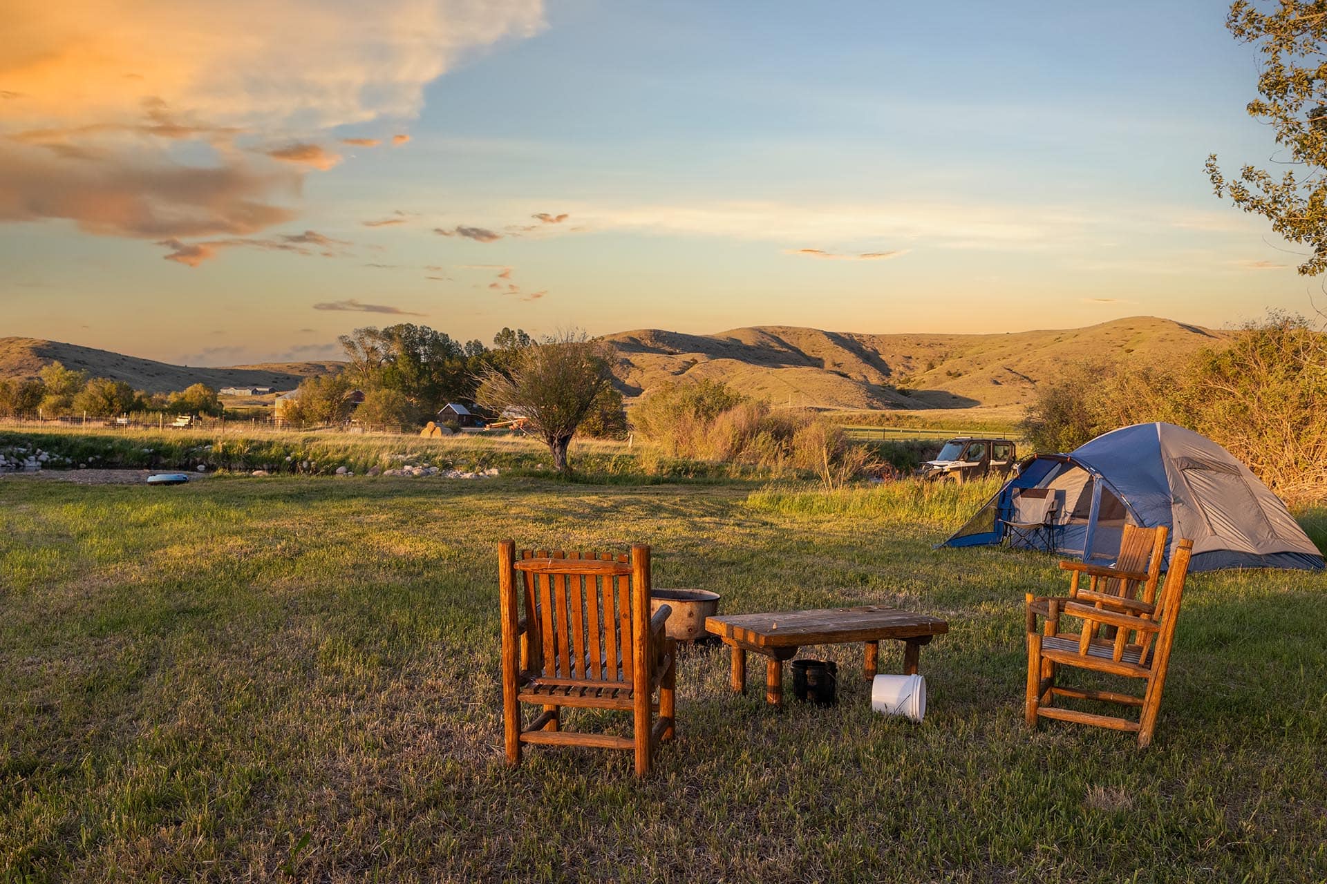 Campsite at sunset with a blue tent, wooden chairs around a fire pit on a grassy field and rolling hills in the distance.