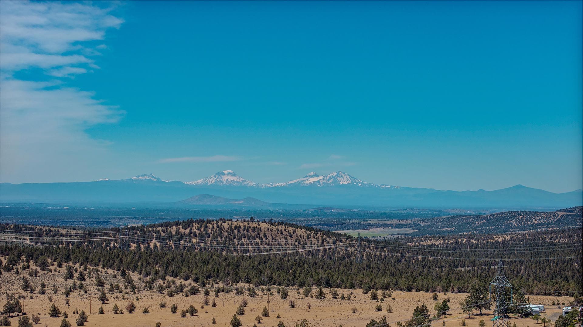 Wide panorama of an arid valley with scattered trees, power lines, and distant snow-capped mountains under a clear blue sky.