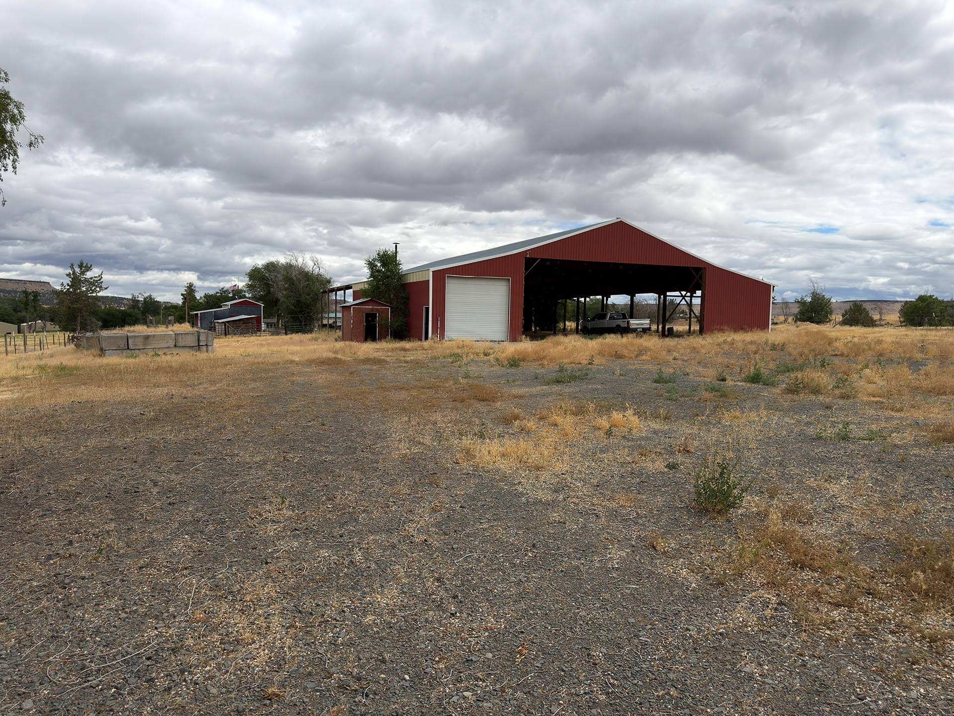 Red metal barn with open front in a dry, rural field under a cloudy sky; a pickup truck inside the barn.