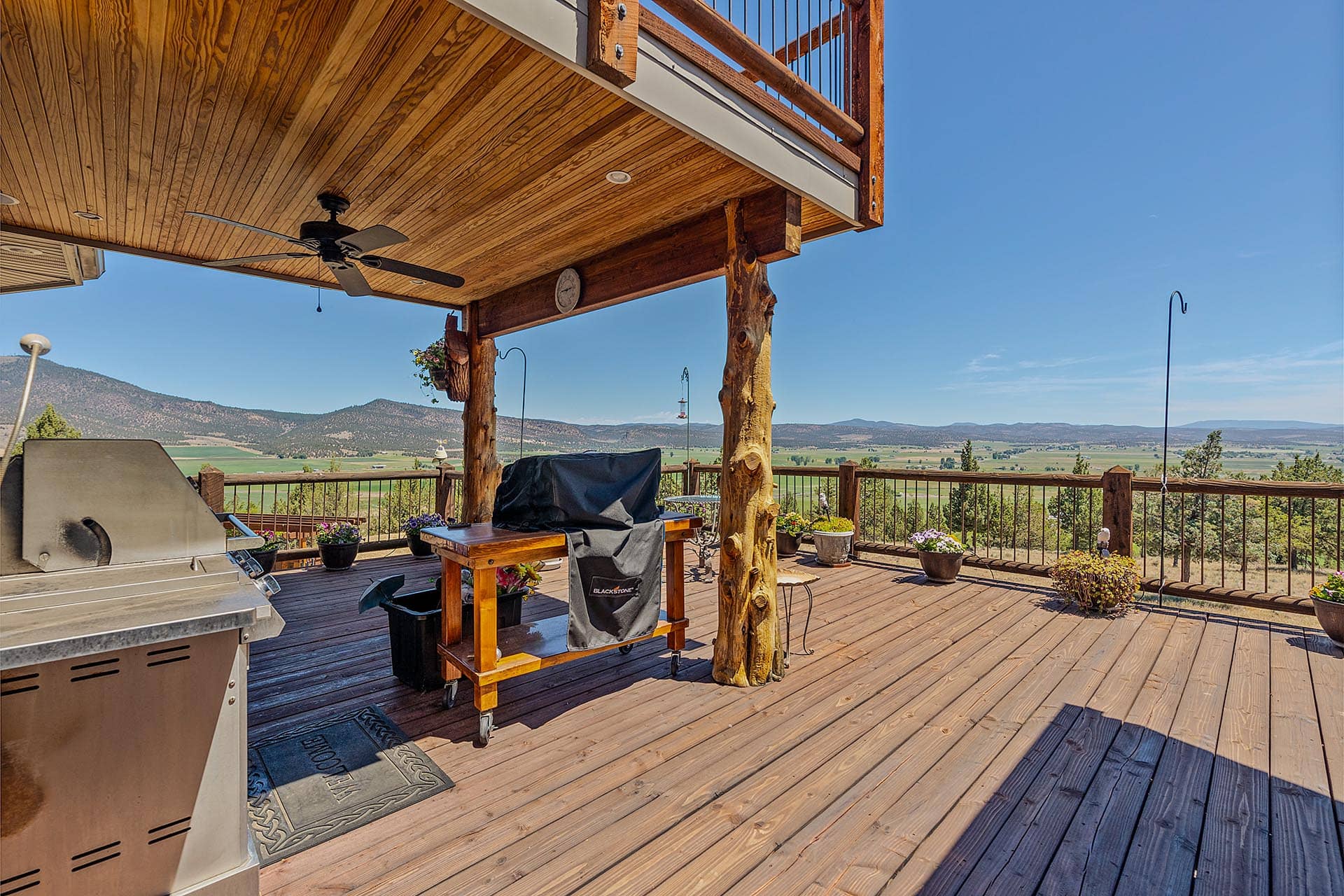 Wooden deck with a grill and a cart under a wooden balcony, overlooking a valley and distant hills against a clear blue sky.