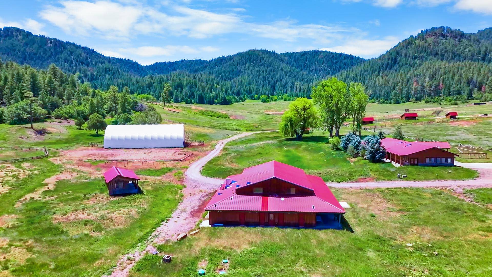 A rural farmscape with red-roofed buildings, a white curved barn, dirt roads, and green hills under a blue sky.