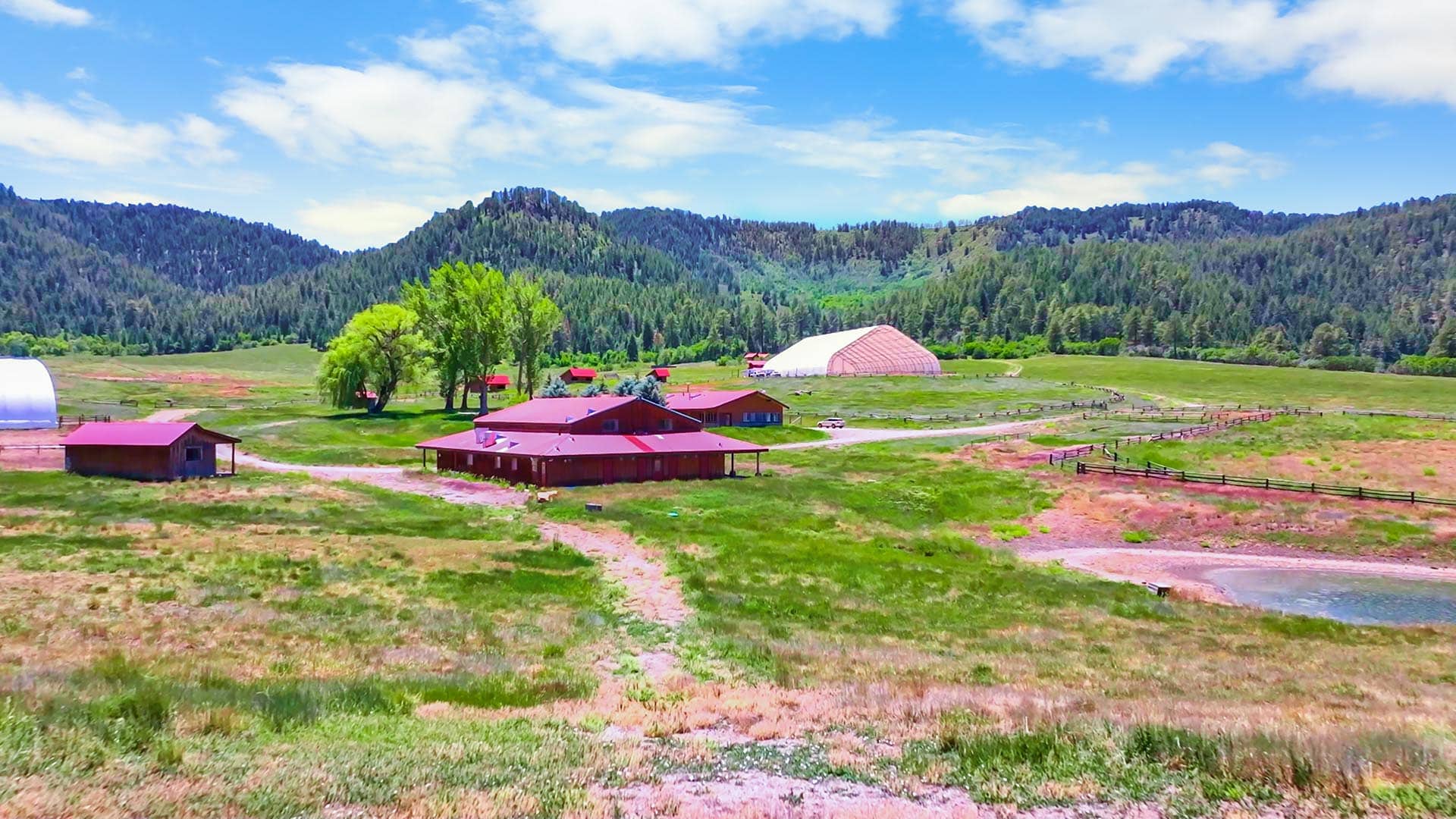 Rural farm scene with red barns, a white arched greenhouse, green fields, and forested hills under a blue sky.