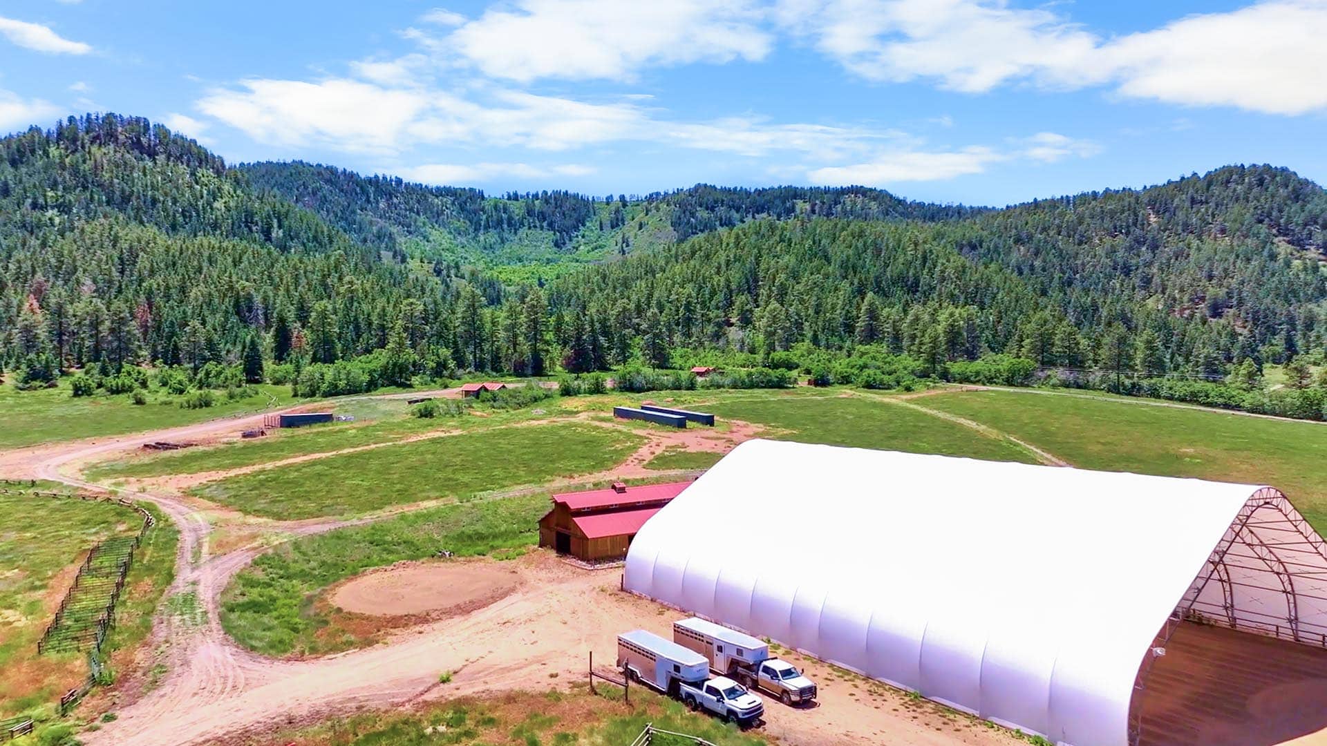 Wide view of a rural farm with a large white greenhouse, red barns, and dirt roads set against forested hills under a blue sky