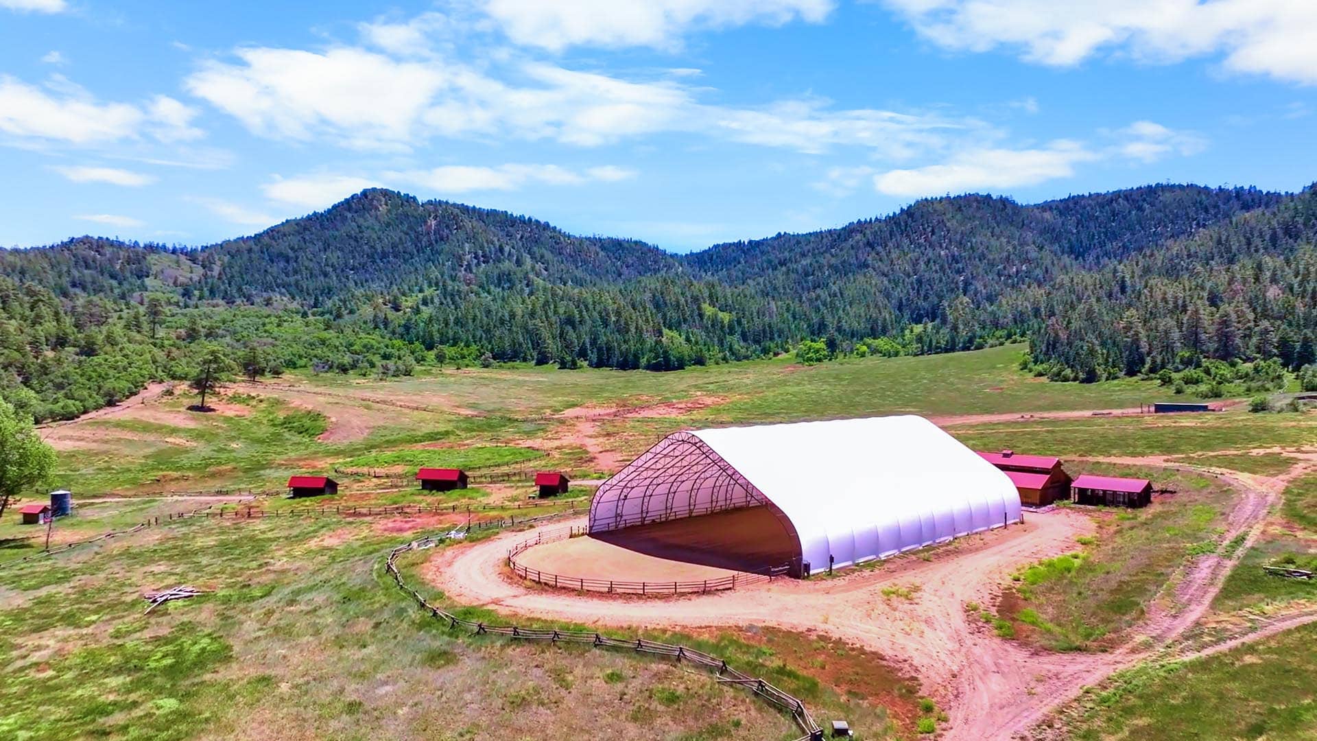 Wide view of a farm with a large white arched shelter, red barns, fences, and green fields, with forested mountains in the background under a blue sky.