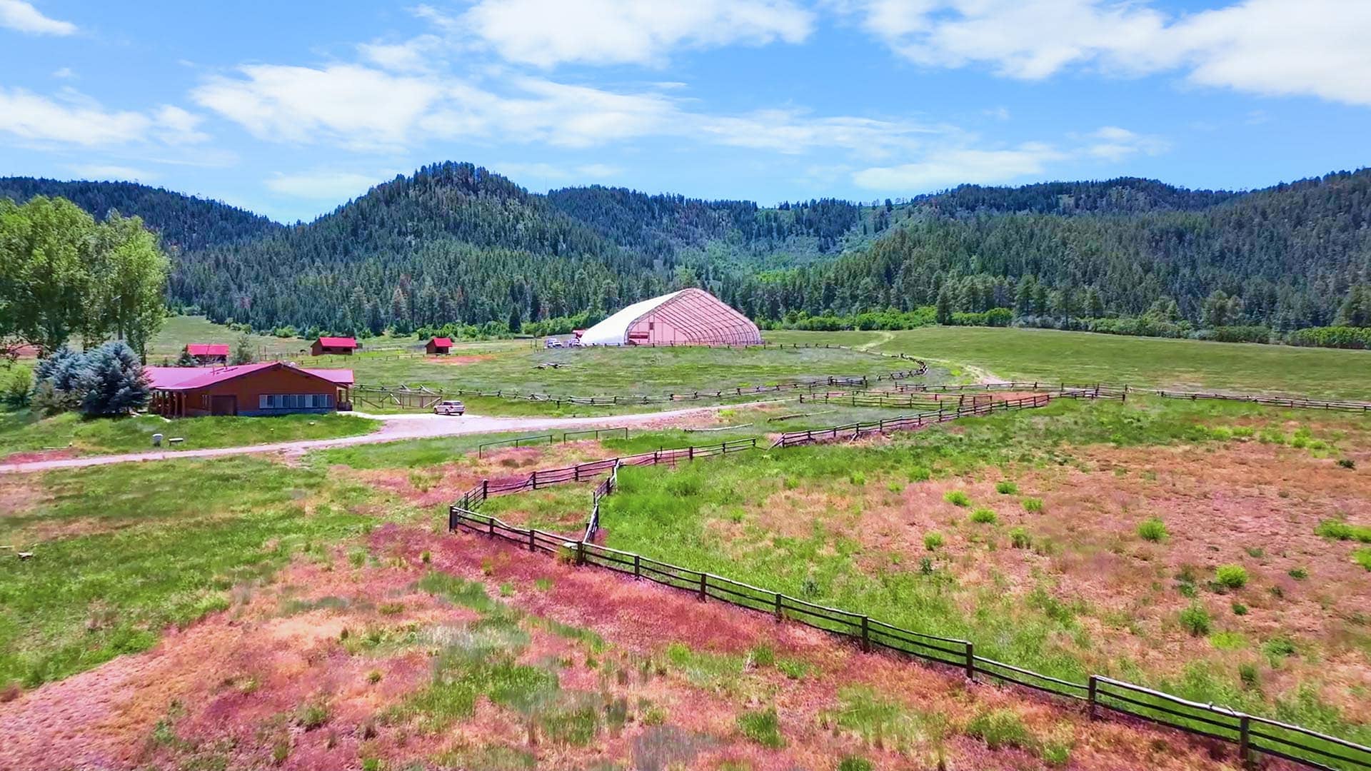 Open countryside with red barn, white-domed greenhouse, scattered small sheds, and winding fences against a mountain backdrop under a blue sky.