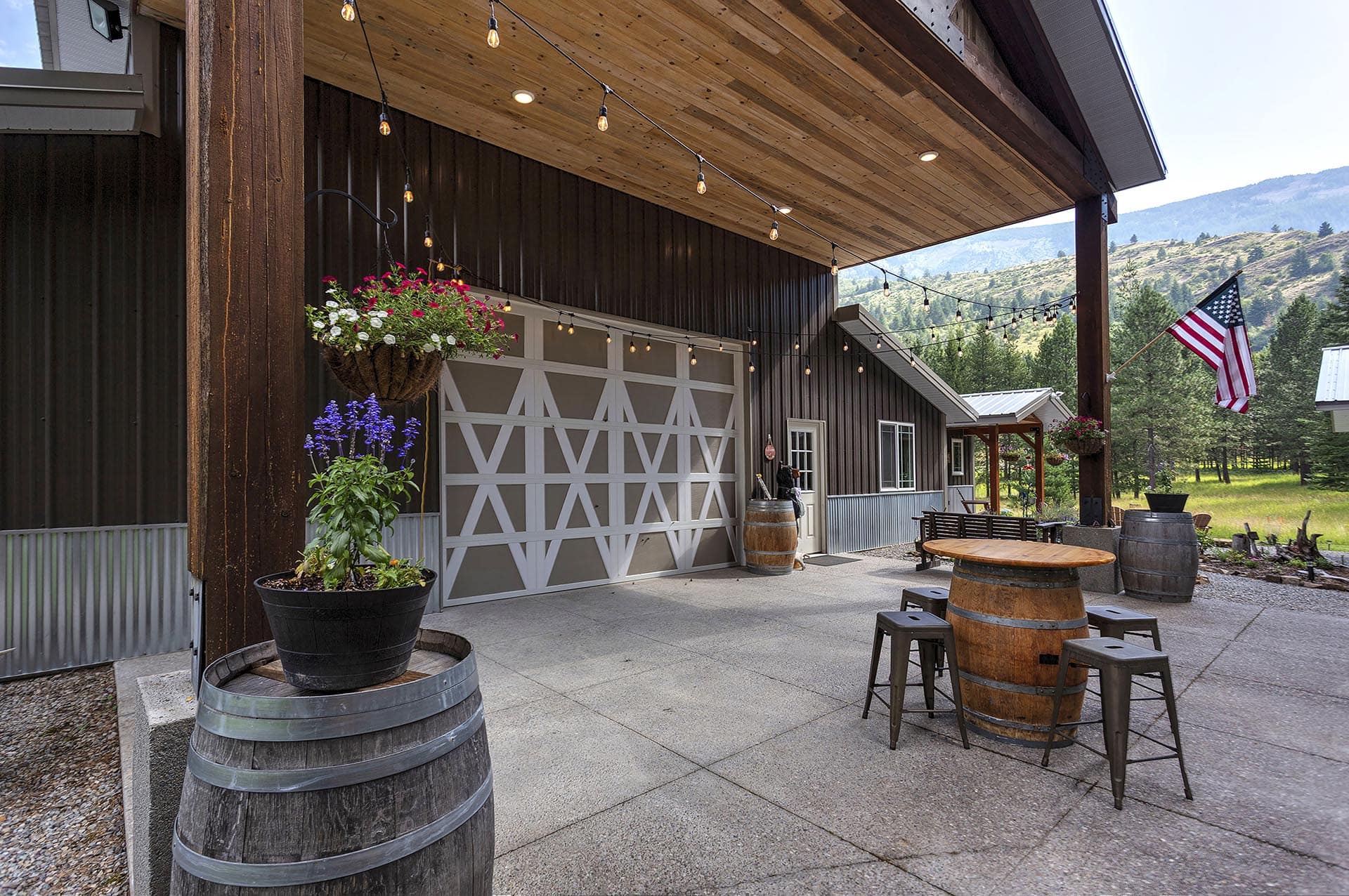 Outdoor patio with string lights, hanging flower baskets, barrel-topped tables, and an American flag against a mountain backdrop.