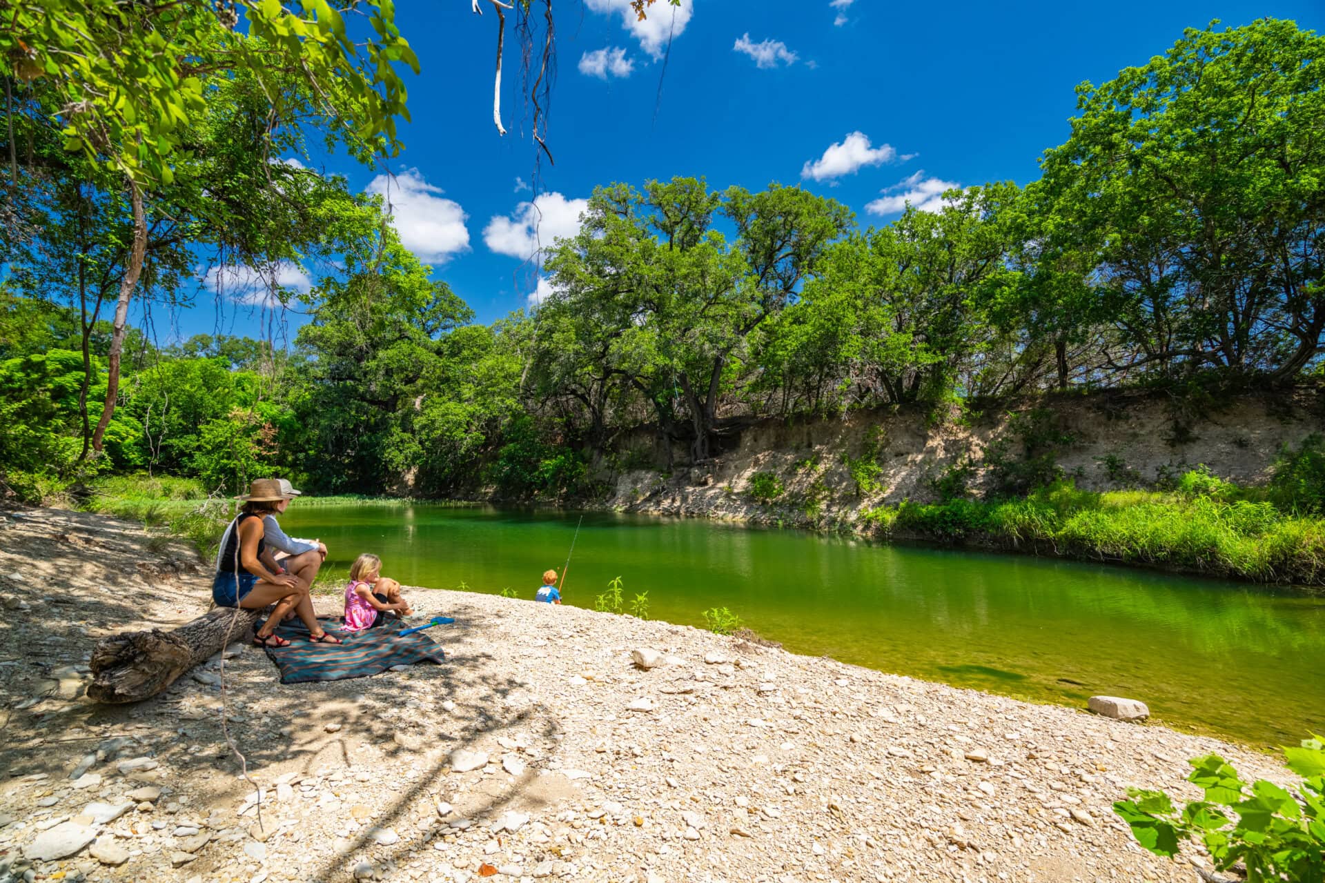 Family sitting on a rocky riverbank fishing under a bright blue sky with green trees around.