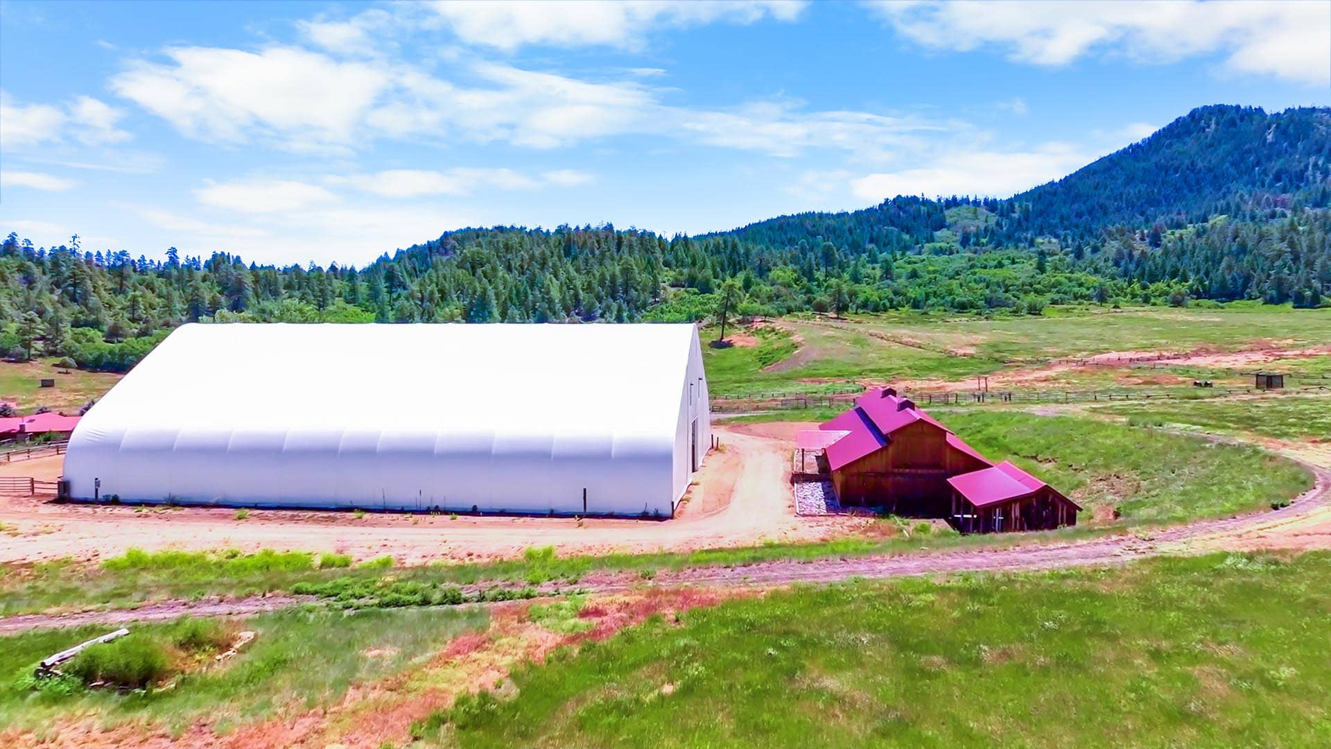 Large white curved-roof storage building and red-roofed barns sit in a grassy rural field with forested hills in the distance under a blue sky.