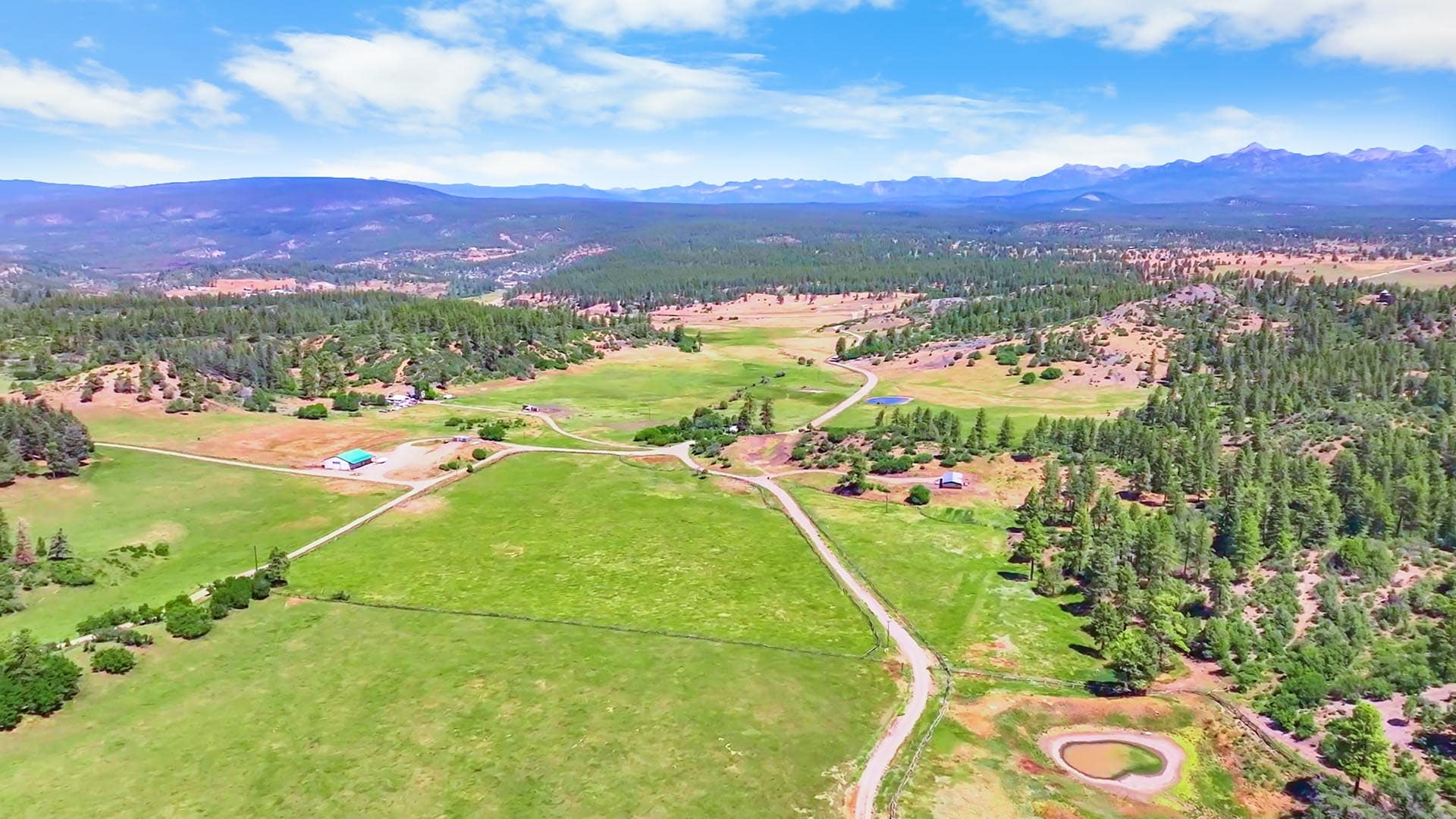 Aerial view of a rural landscape with green fields, winding roads, scattered buildings, and forested hills under a blue sky with distant mountains